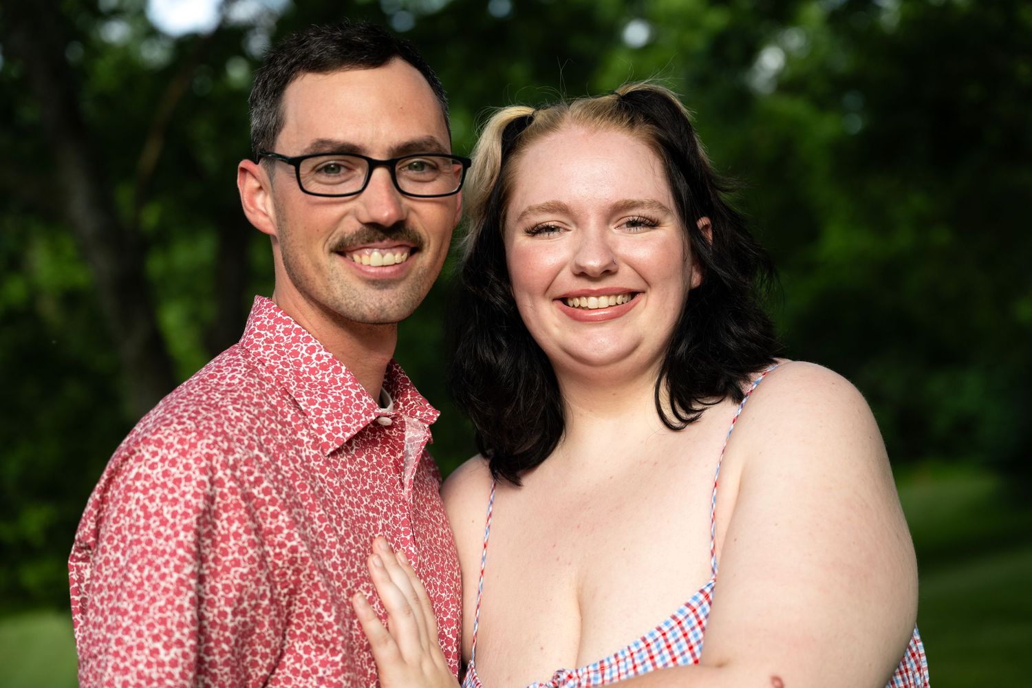 portrait of a Smiling couple poses outdoors in a lush green park, man in pink floral shirt and woman in striped dress.