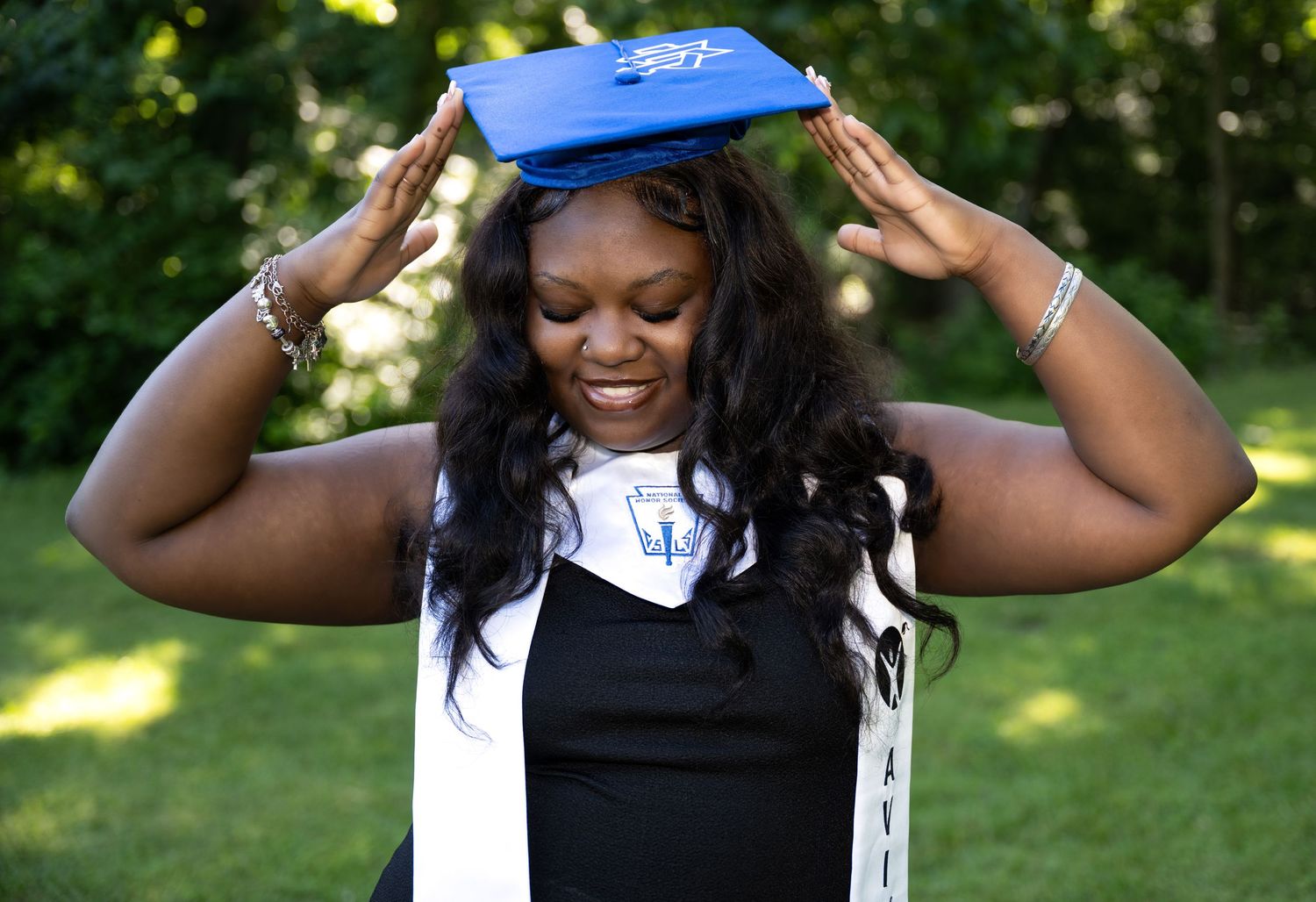 Happy graduate adjusting blue graduation cap while smiling in a green outdoor park setting.