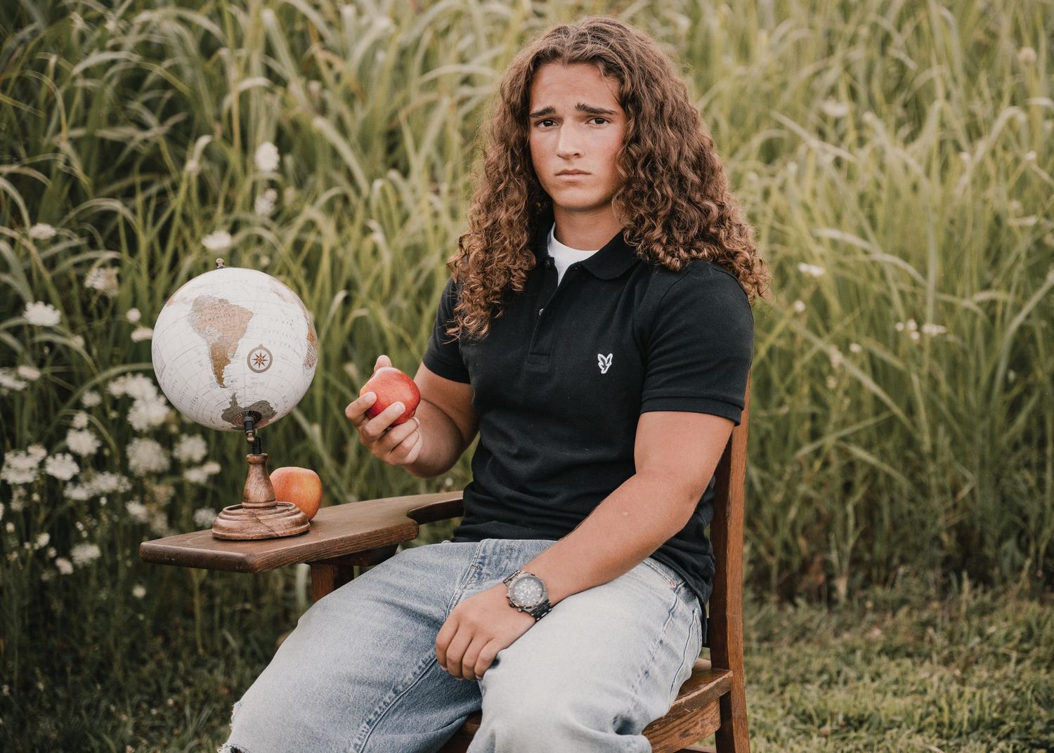 portrait of a Young man with curly hair sits on chair holding apple near globe outdoors.