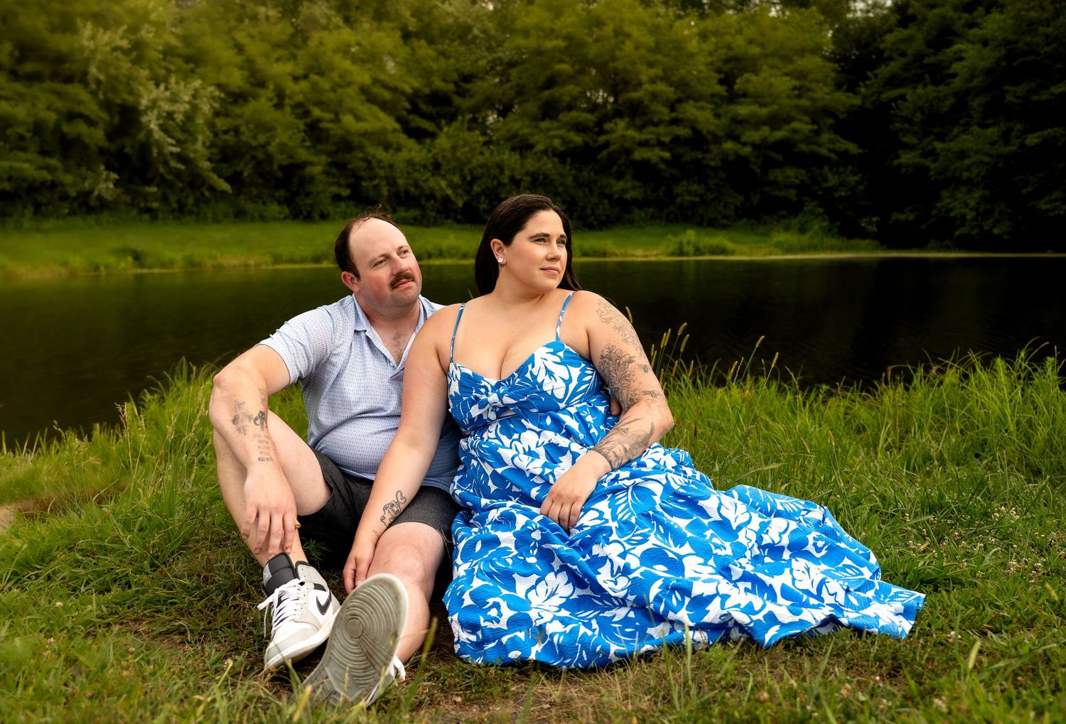 lifestyle portrait of a Couple sitting on grass by a lake, woman in blue floral dress, man in white shirt, surrounded by lush green trees.