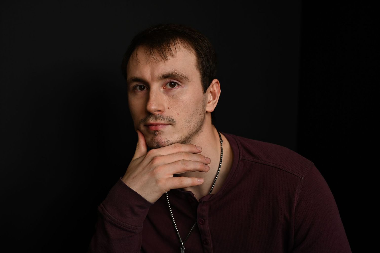 A thoughtful young man in a maroon shirt rests his hand on his chin against a dark studio background.