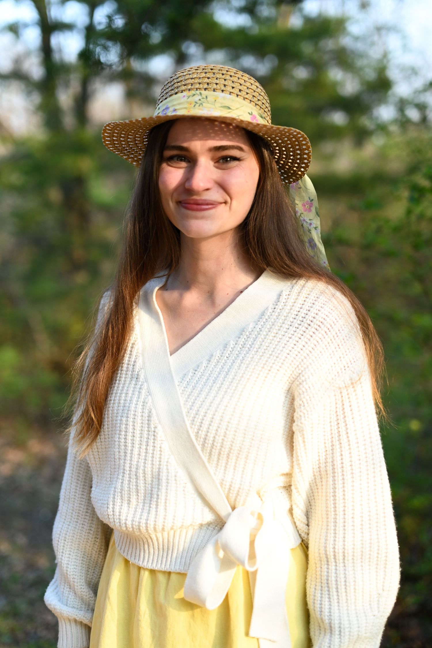 Smiling young woman in cream knit wrap sweater and straw hat in a sunny garden.