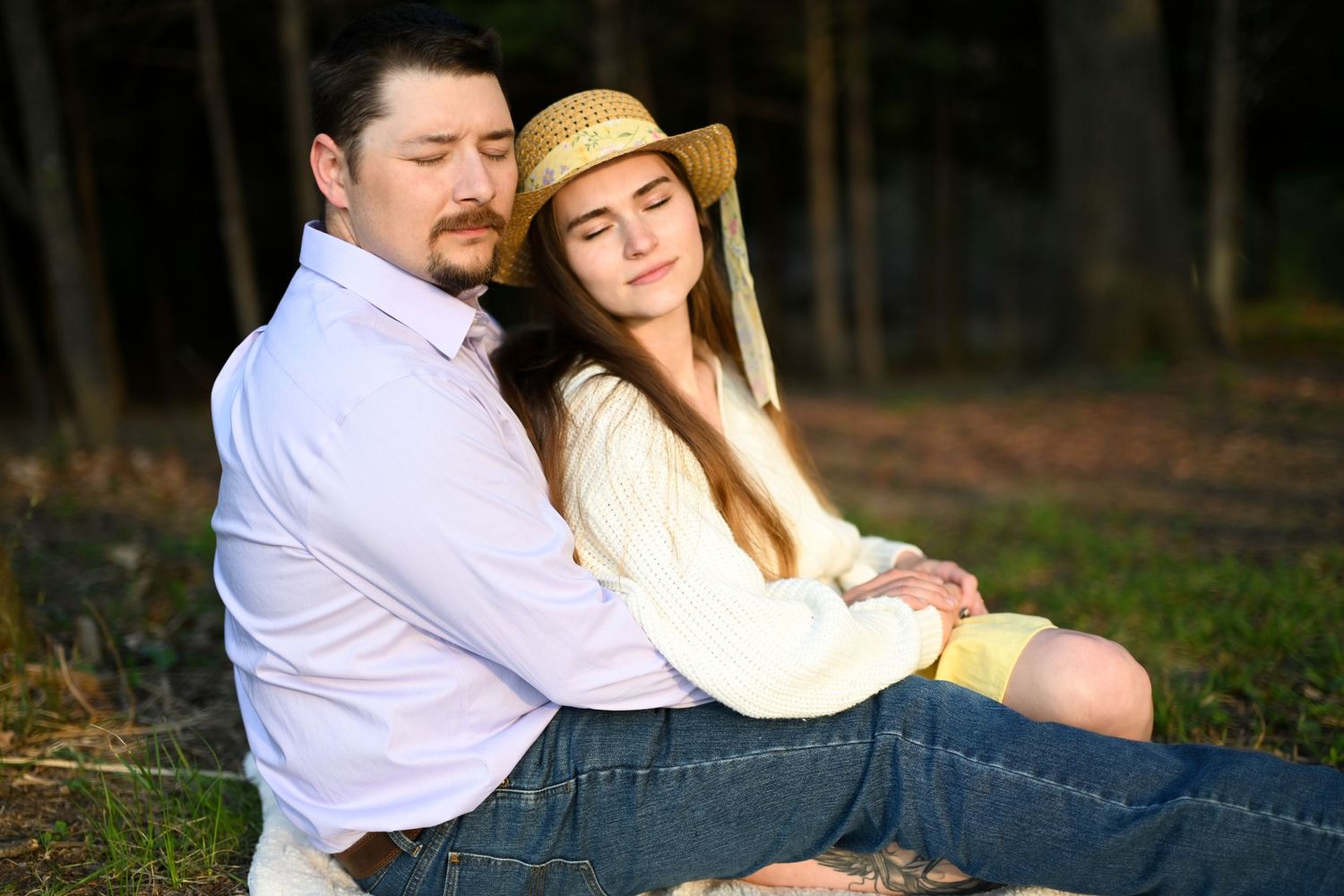 A couple sits peacefully together in a sunlit forest, woman in yellow dress leaning on mans shoulder.
