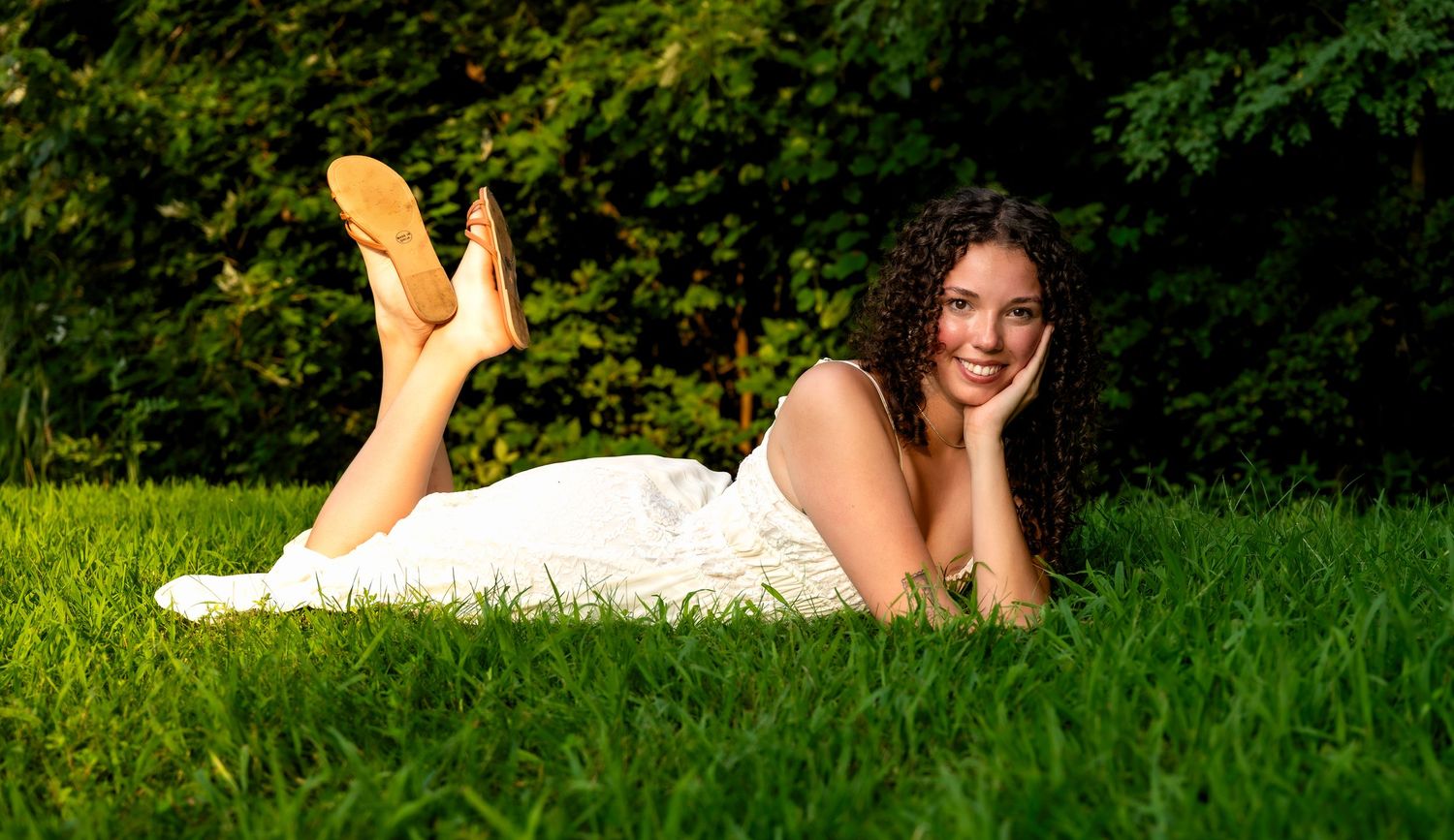 lifestyle portrait of a Young woman in white dress lying on lush green grass, smiling with hand on cheek, wearing tan shoes.