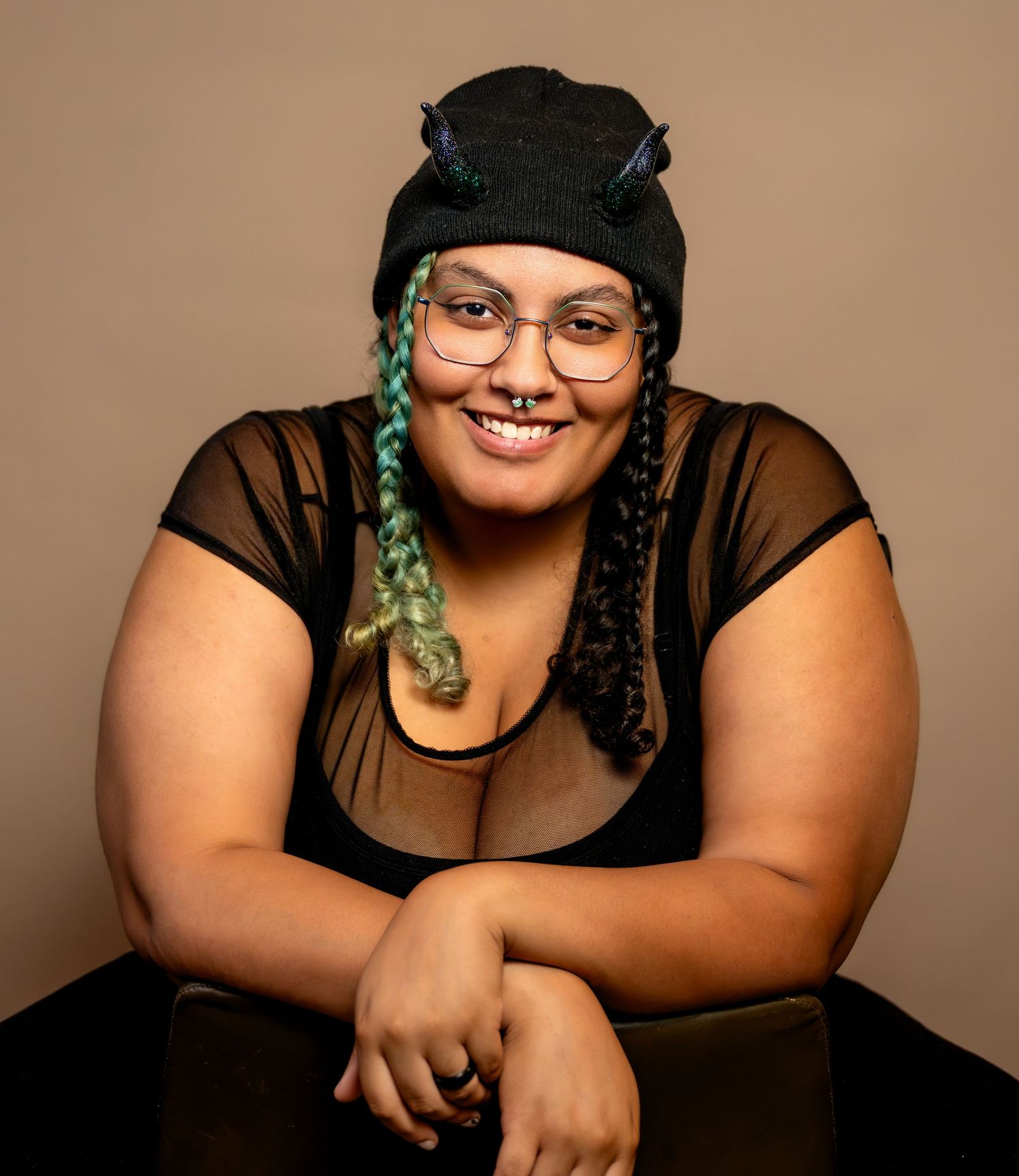 Smiling young woman with braids, glasses, and black hat poses confidently in a stylish black top.