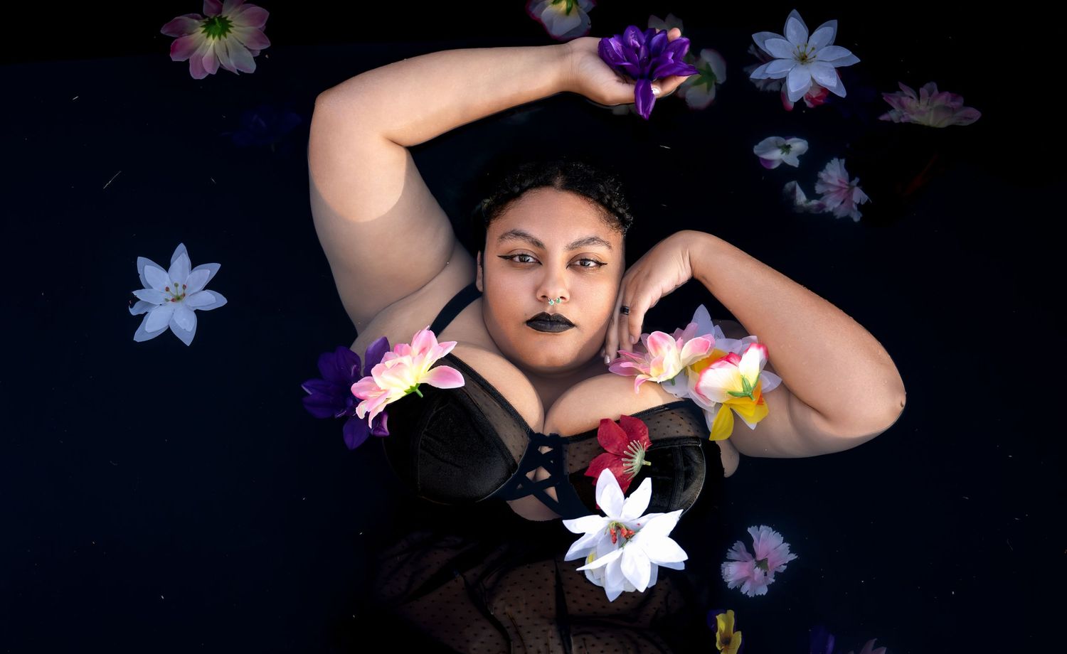 Woman with dark lipstick floating in water surrounded by colorful flowers in artistic portrait photography.