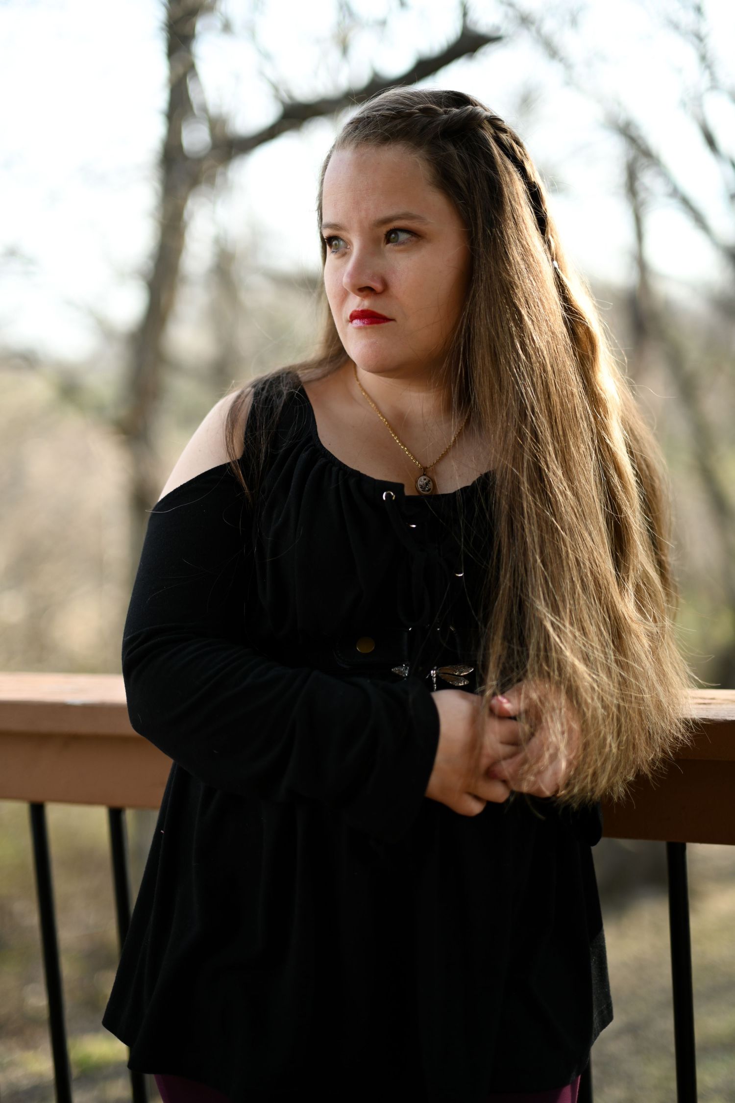 Young woman with long hair in black off-shoulder top leaning on outdoor deck railing.