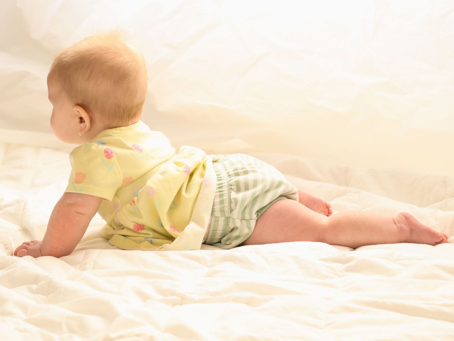 Baby in yellow romper lying on stomach looking sideways on a white blanket.