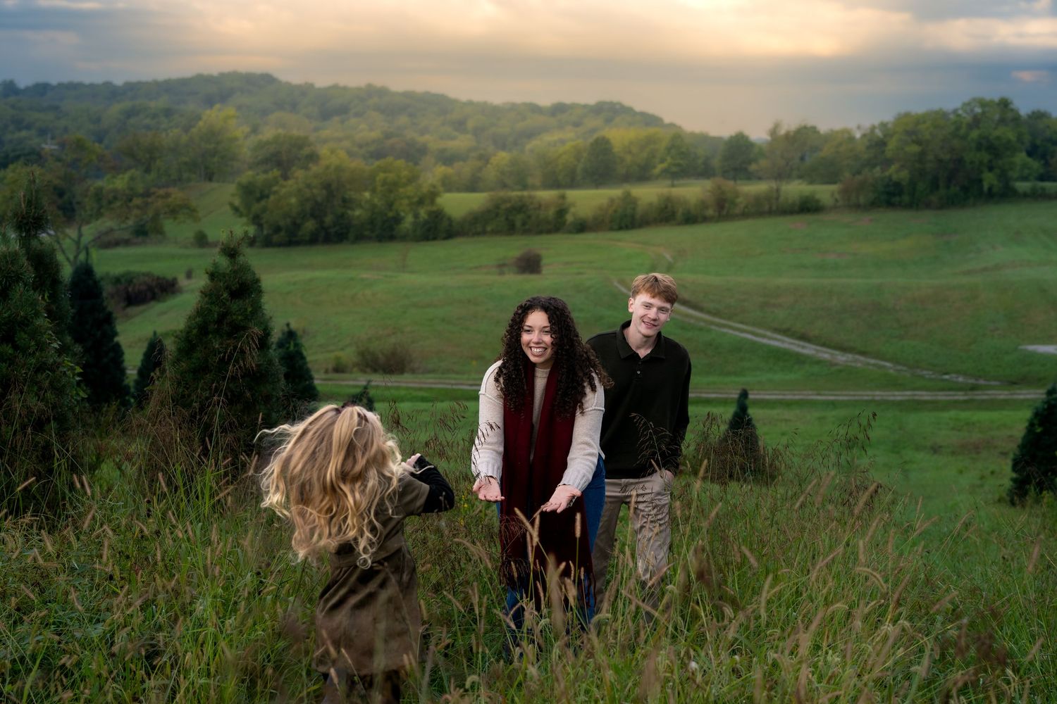 Two teens and a child play in a lush green hillside meadow at dusk with rolling farmland in the background.