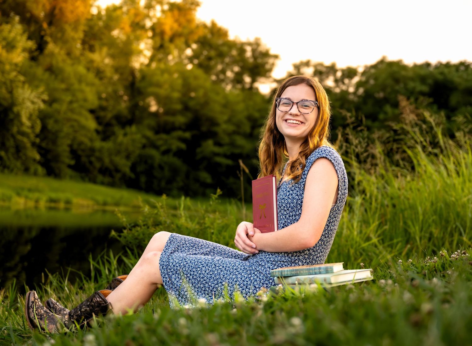 lifestyle portrait of a Young woman with glasses sits in green grass holding books, smiling outdoors at golden hour near a pond.