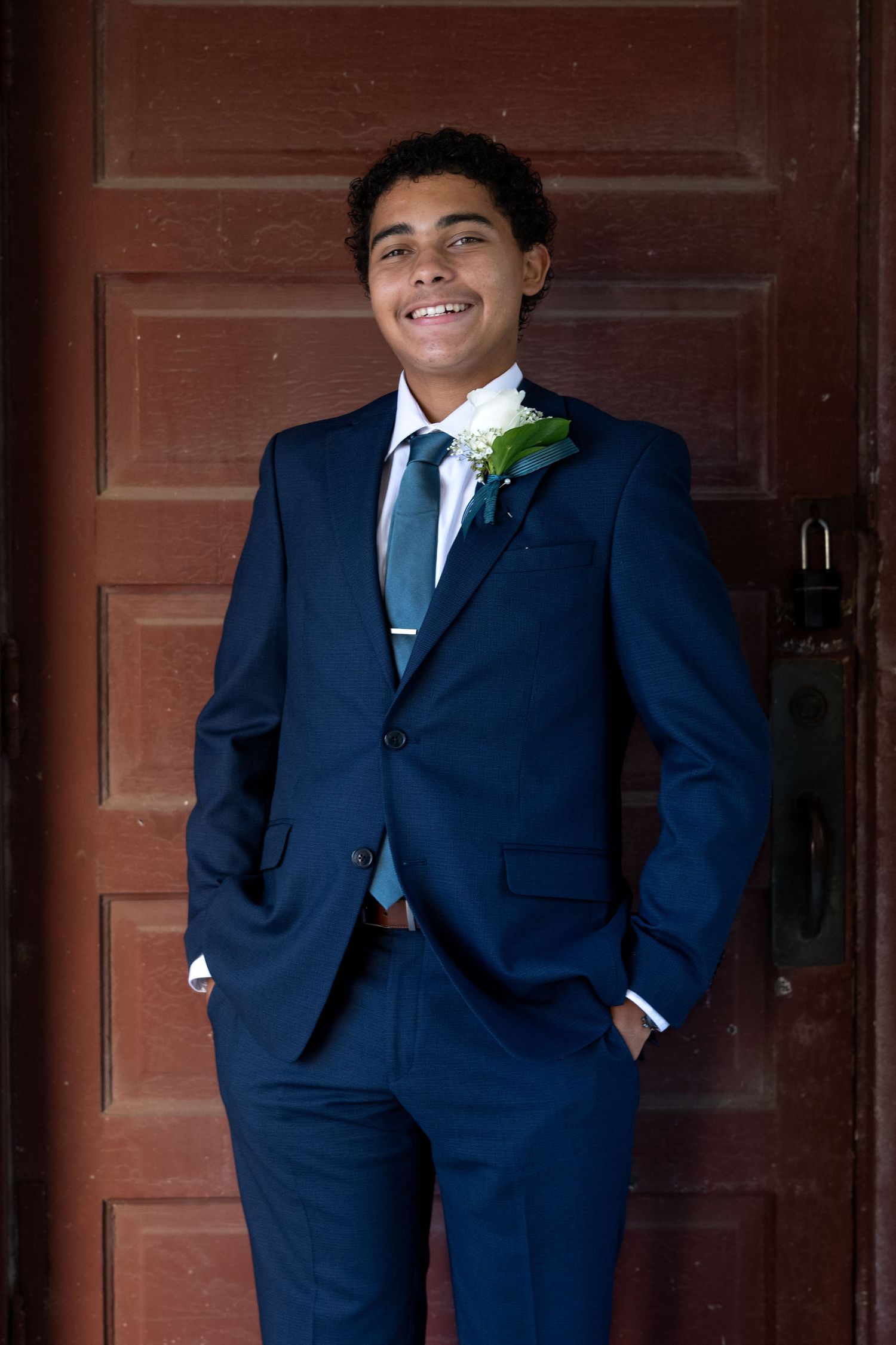 Smiling young man in navy blue suit with boutonniere and teal tie poses in front of rustic red wooden door.
