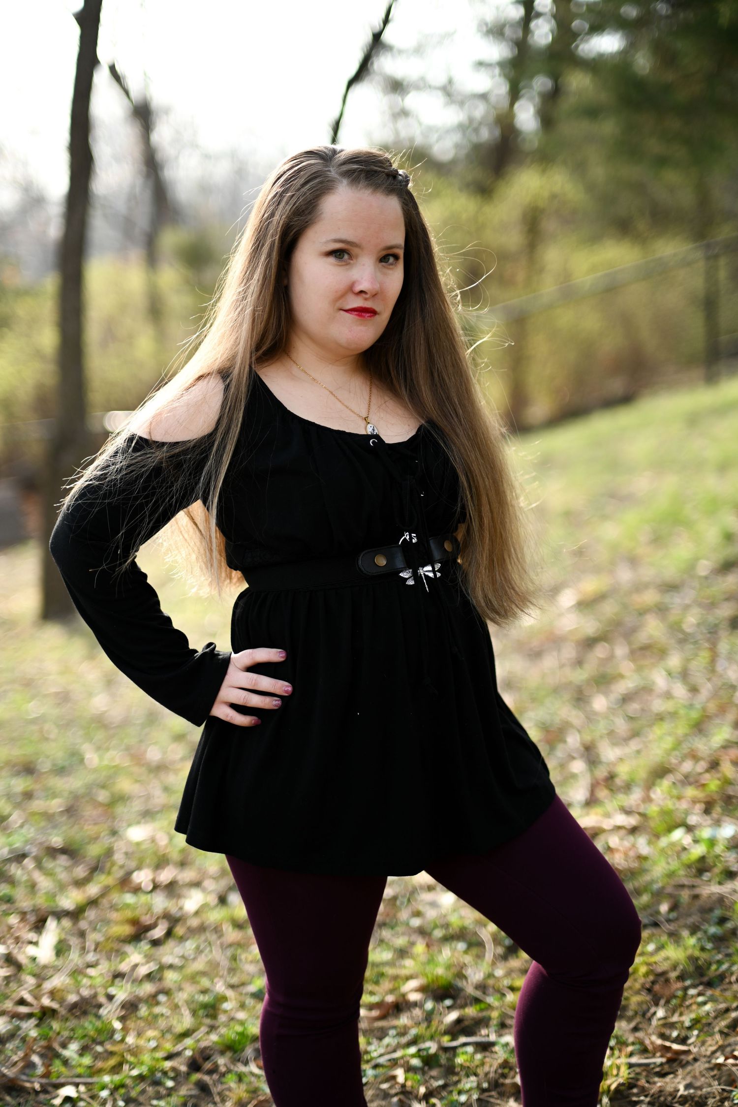 Young woman in black top and purple leggings posing outdoors in wooded setting.