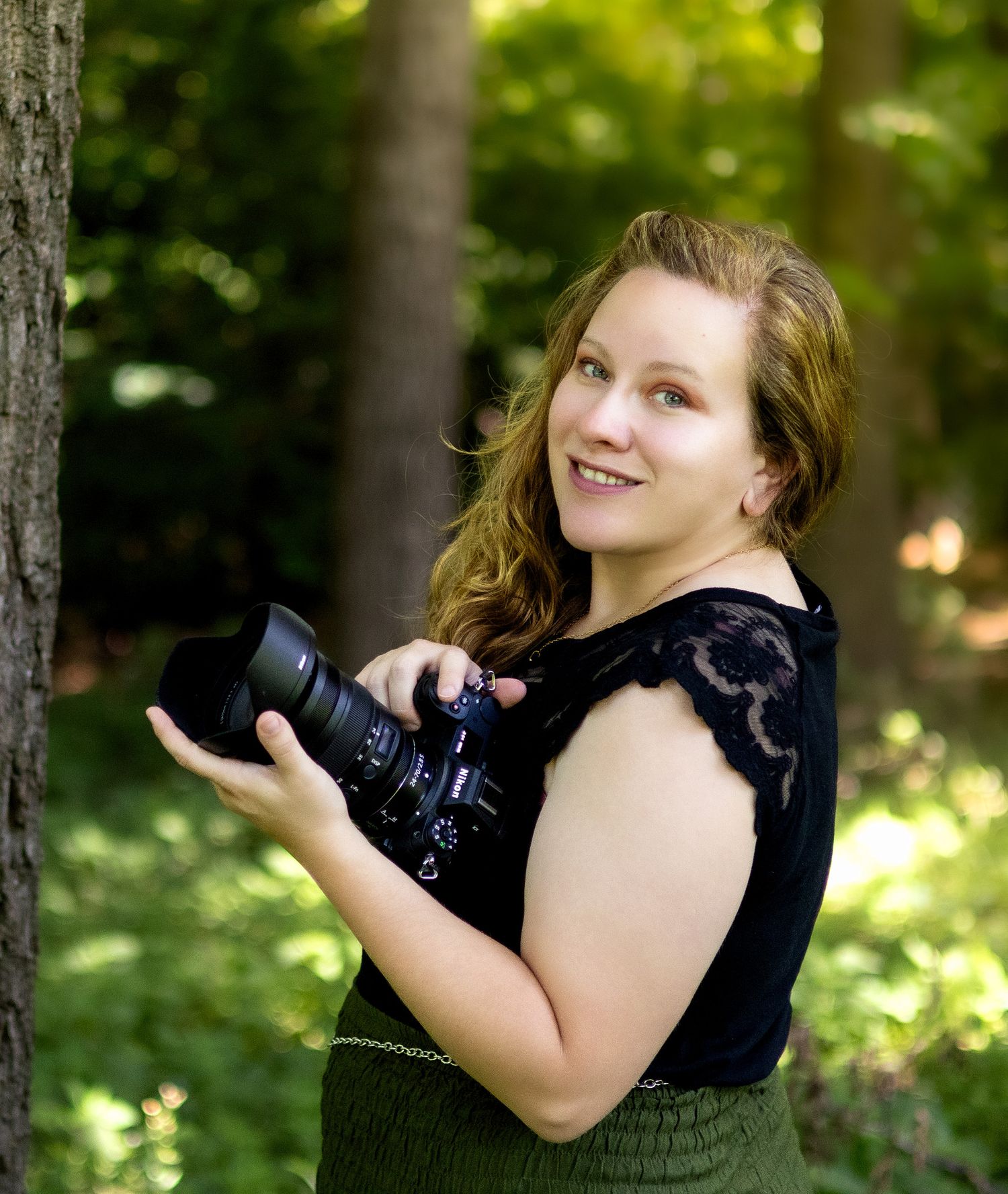 Young woman with curly hair holding a DSLR camera, smiling outdoors in a lush green forest setting.