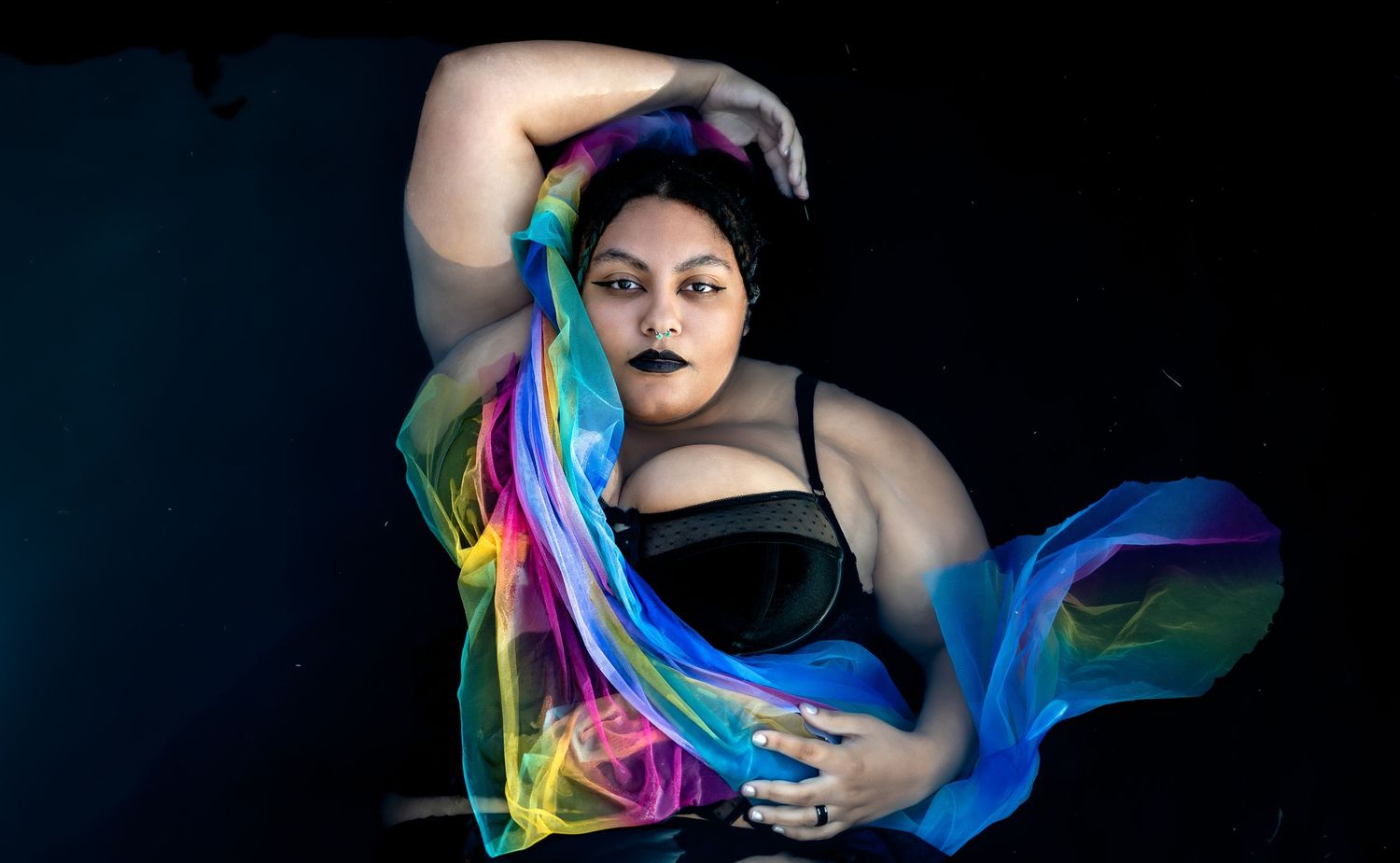 Plus-size woman posing underwater with vibrant rainbow fabric flowing around her in a dark pool.