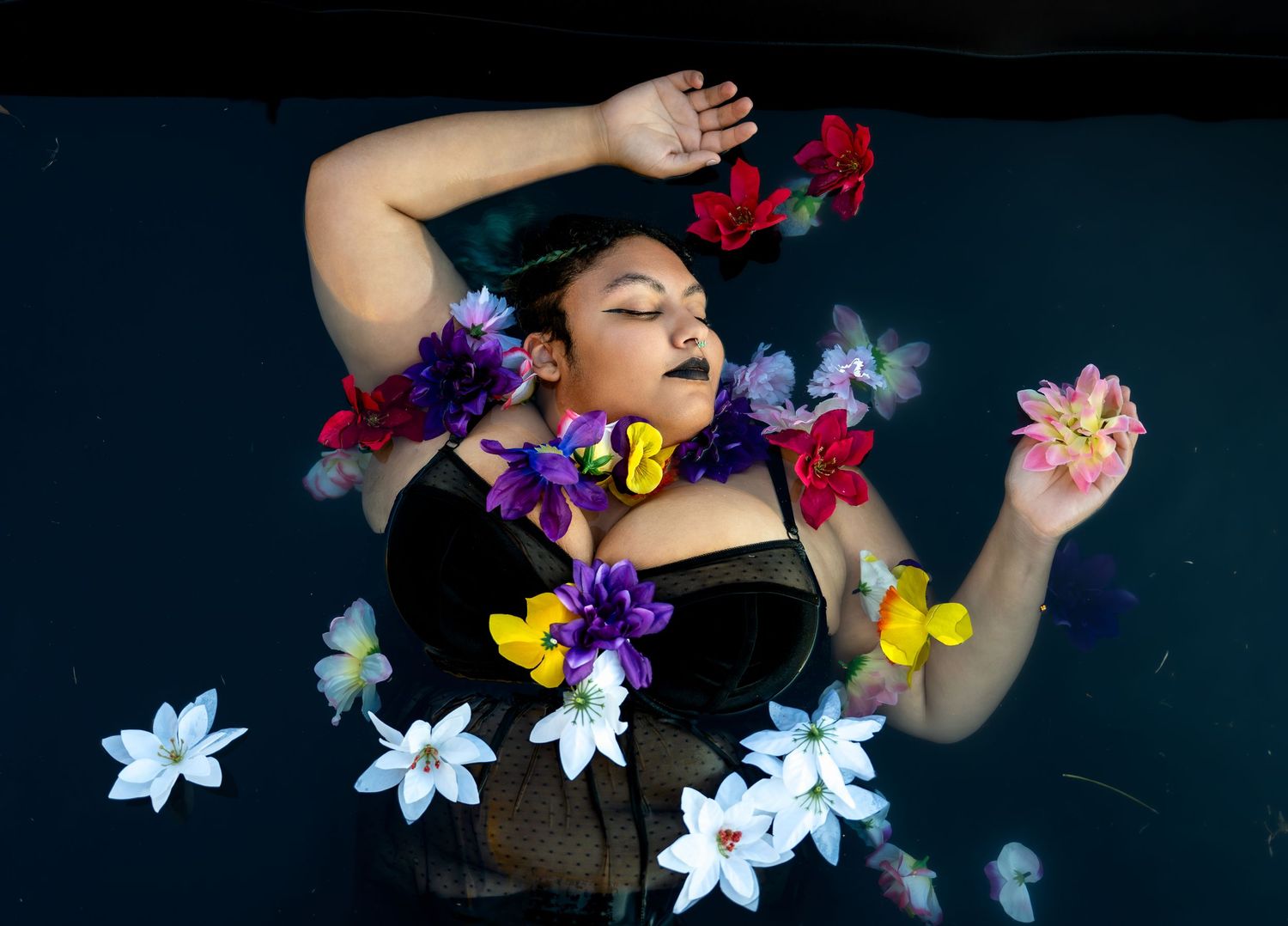 Woman floating in water surrounded by colorful flowers in a serene and artistic floral bath photography session.