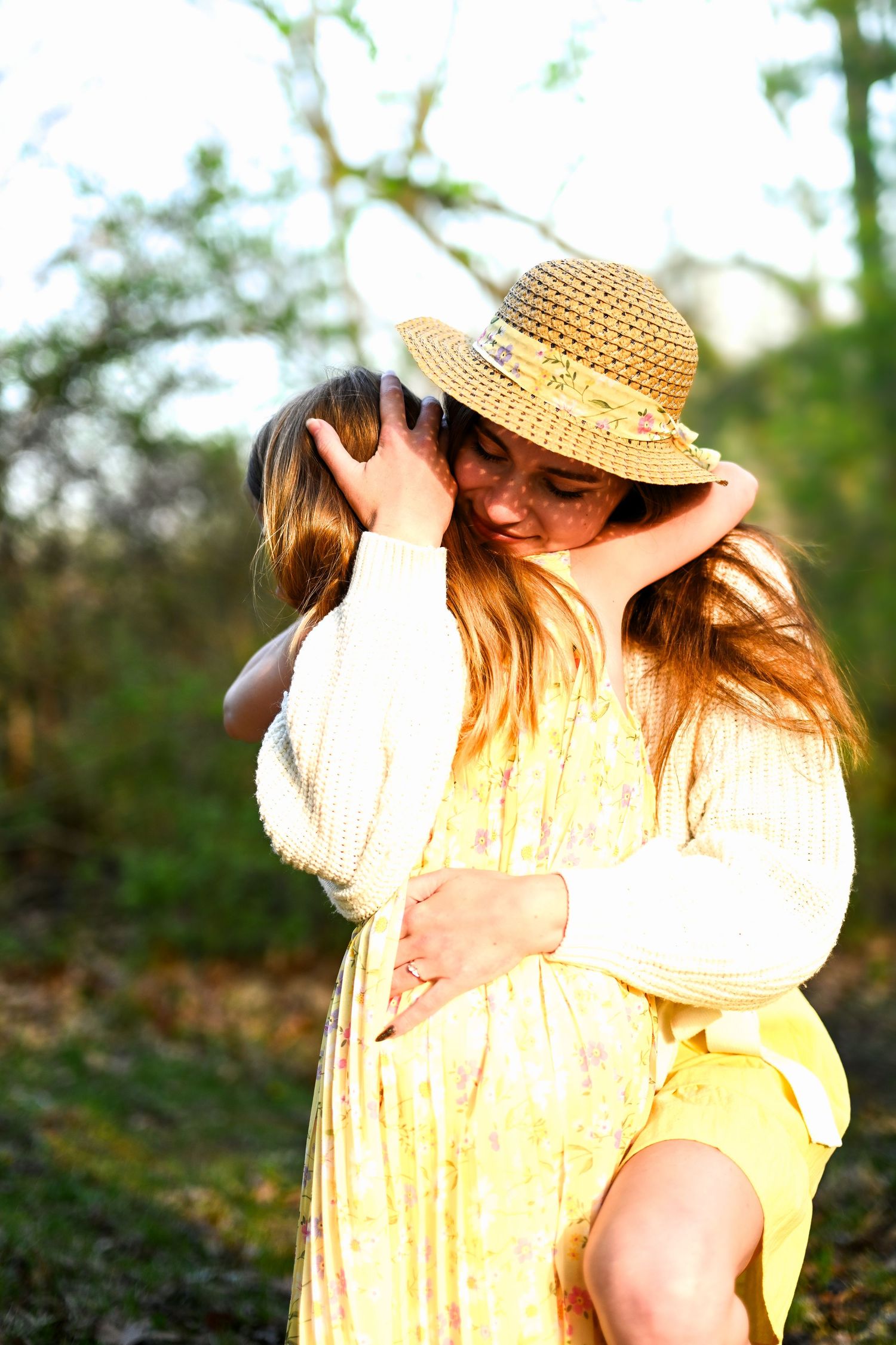 A woman in a yellow floral dress and straw hat holds a child outdoors in warm golden sunlight.