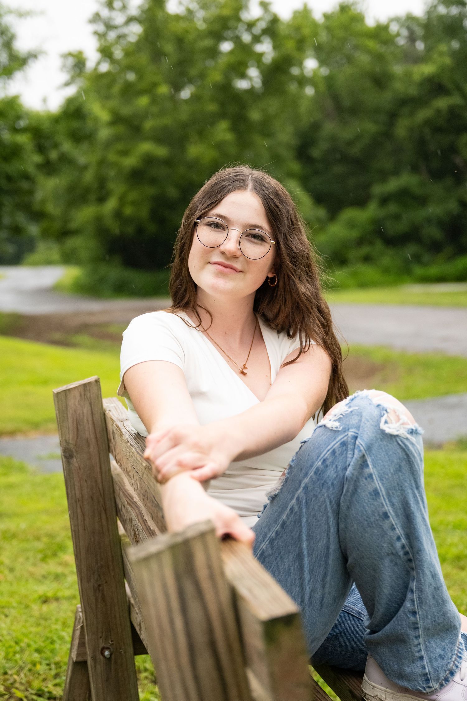portrait of a Young woman in glasses and jeans relaxing on wooden park bench with green trees behind her.