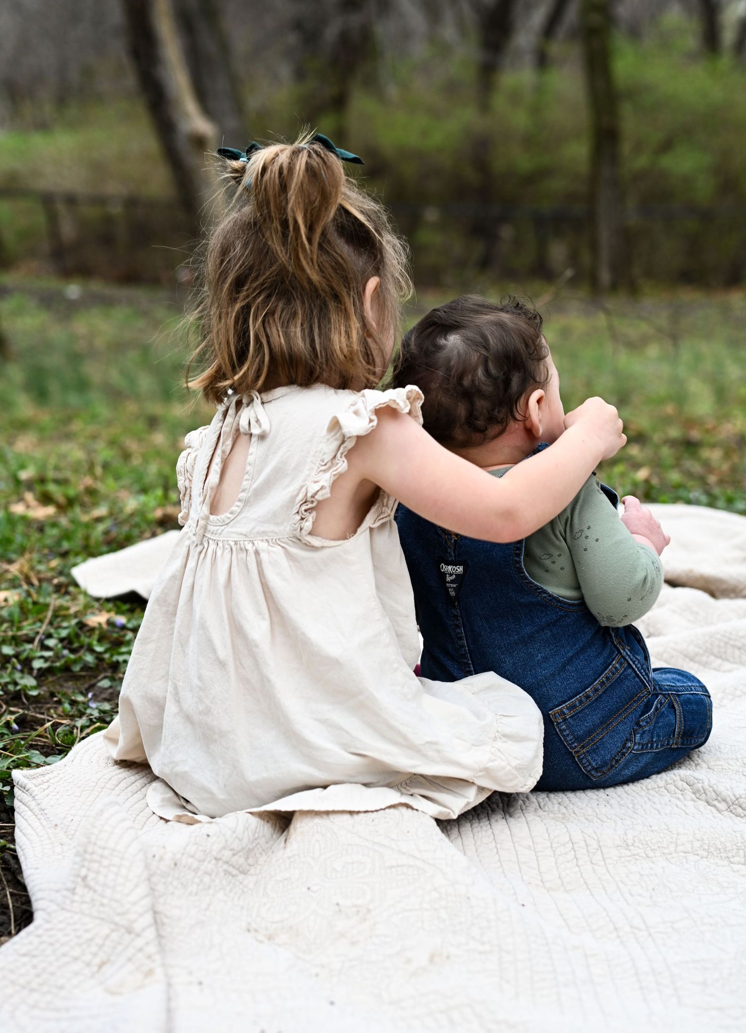 Two young siblings sitting together on white blanket outdoors in wooded park setting from behind.