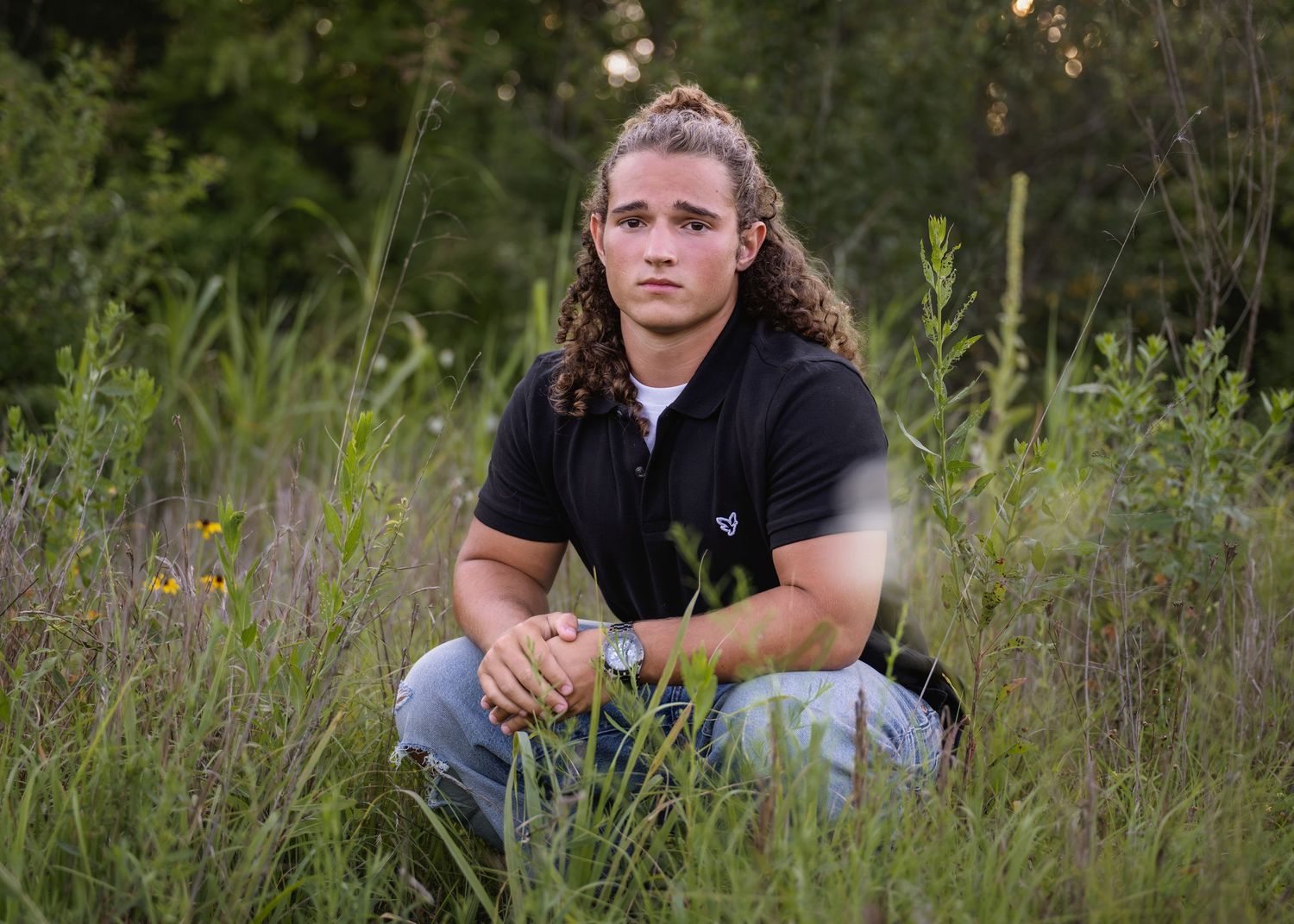 portrait of a young man with curly hair crouches in lush green field wearing black polo.