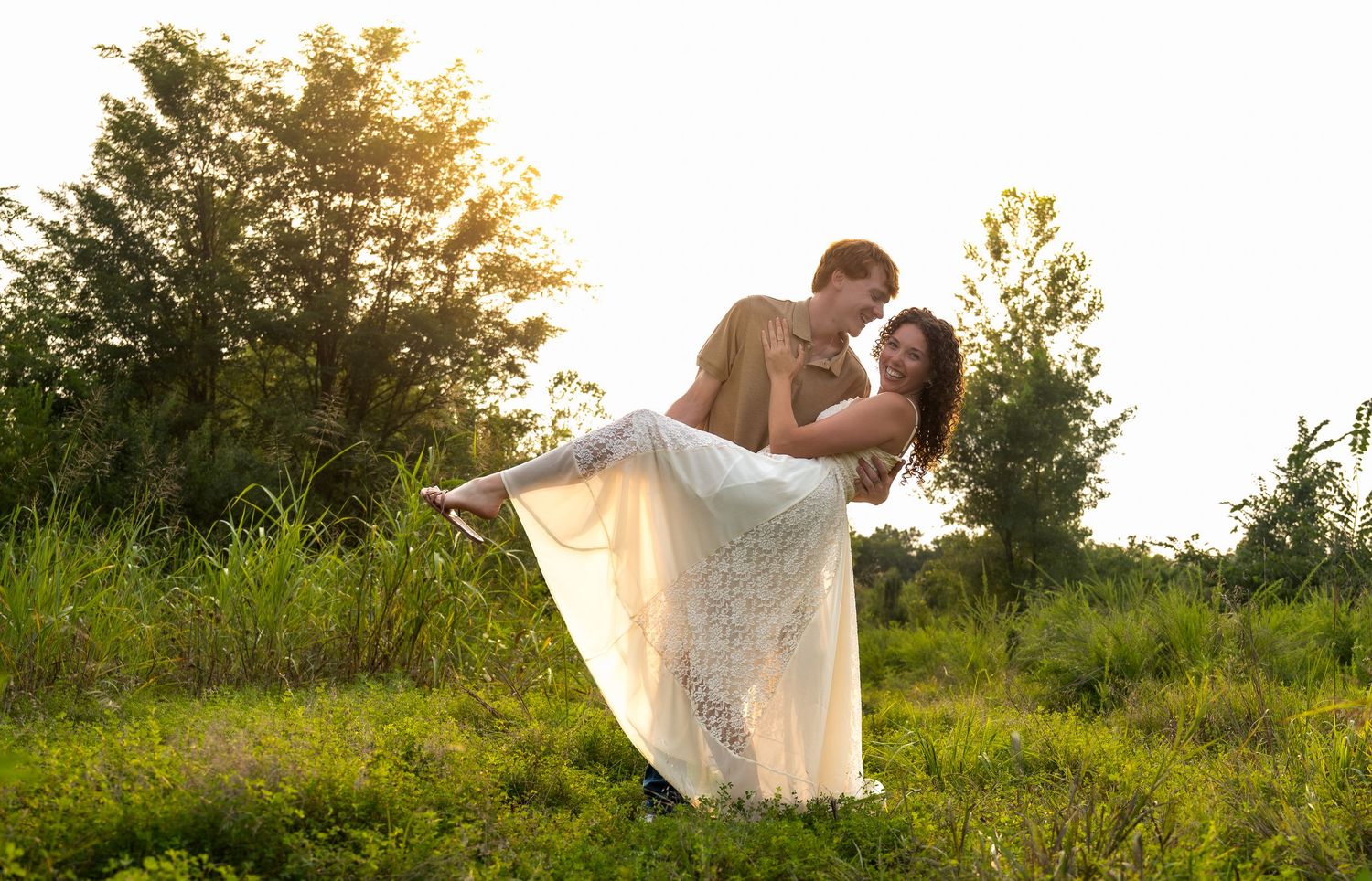 lifestyle portrait of a man lifting wife in white lace dress in sunlit green meadow during golden hour outdoor wedding.