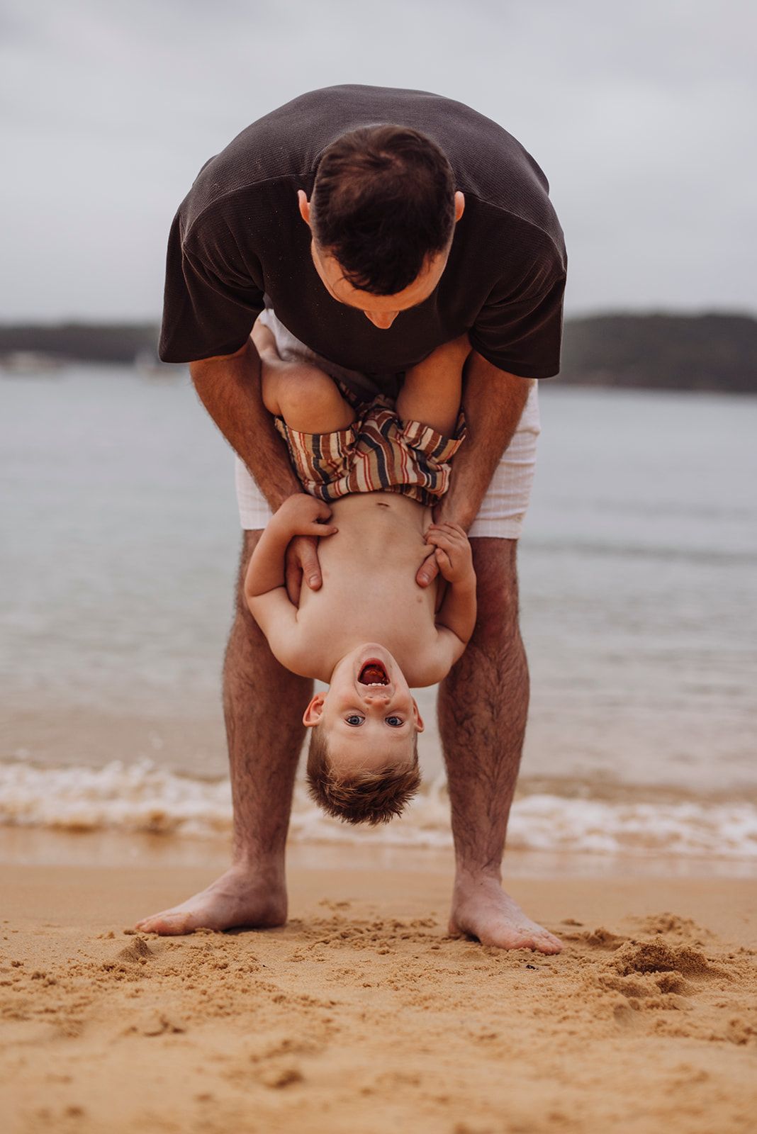 Watsons Bay family photography of father playfully holding child upside down on the sand, IVF success family