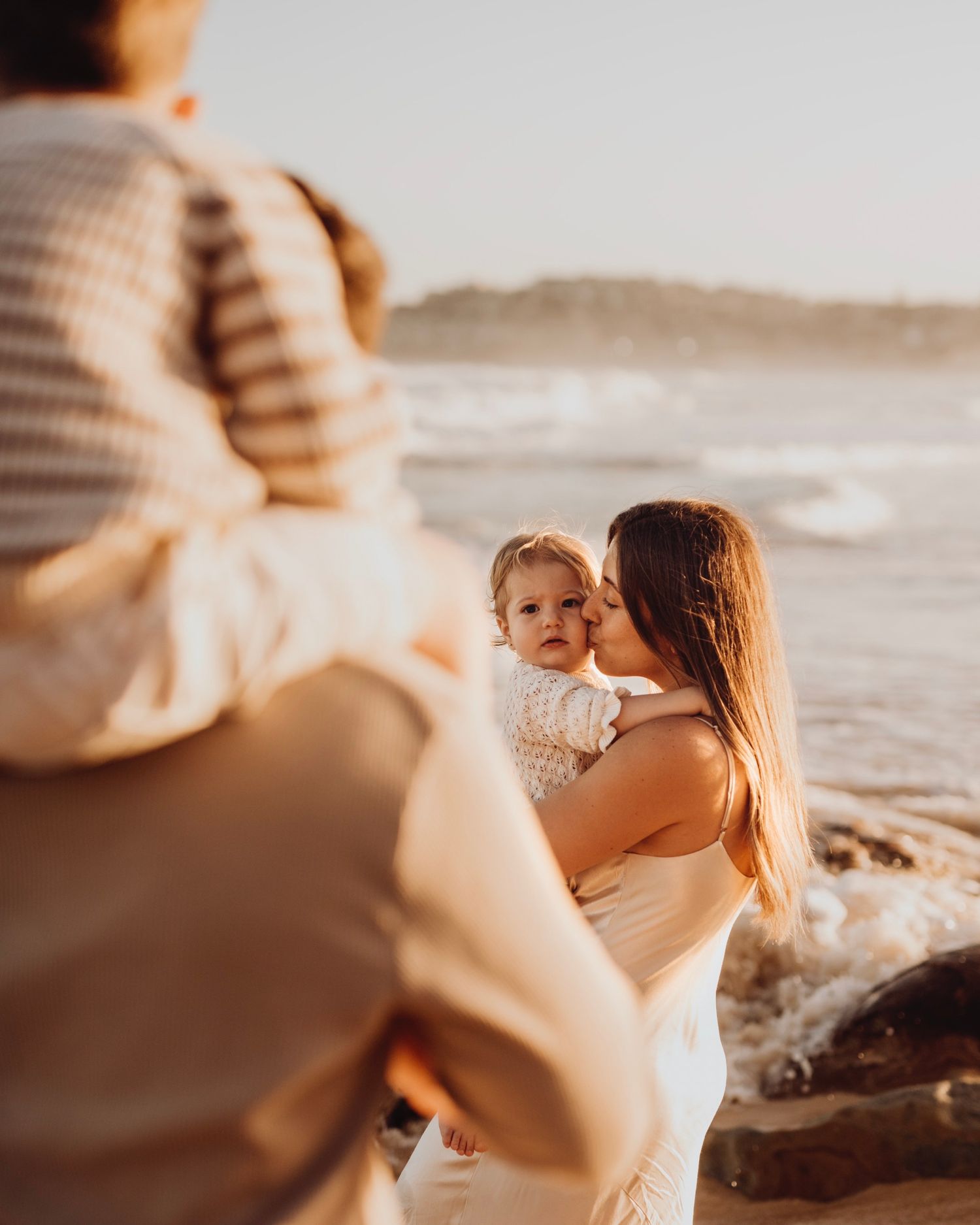 Northern Beaches family photography of mother holding child while father carries another on shoulders by the ocean