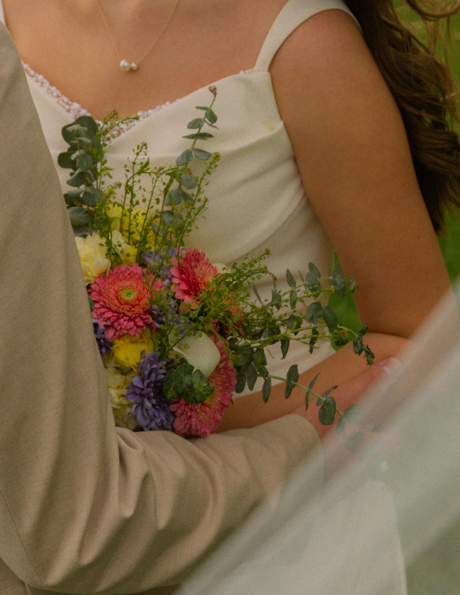 Close-up of bride holding a colorful bouquet with pink daisies and eucalyptus in white dress.