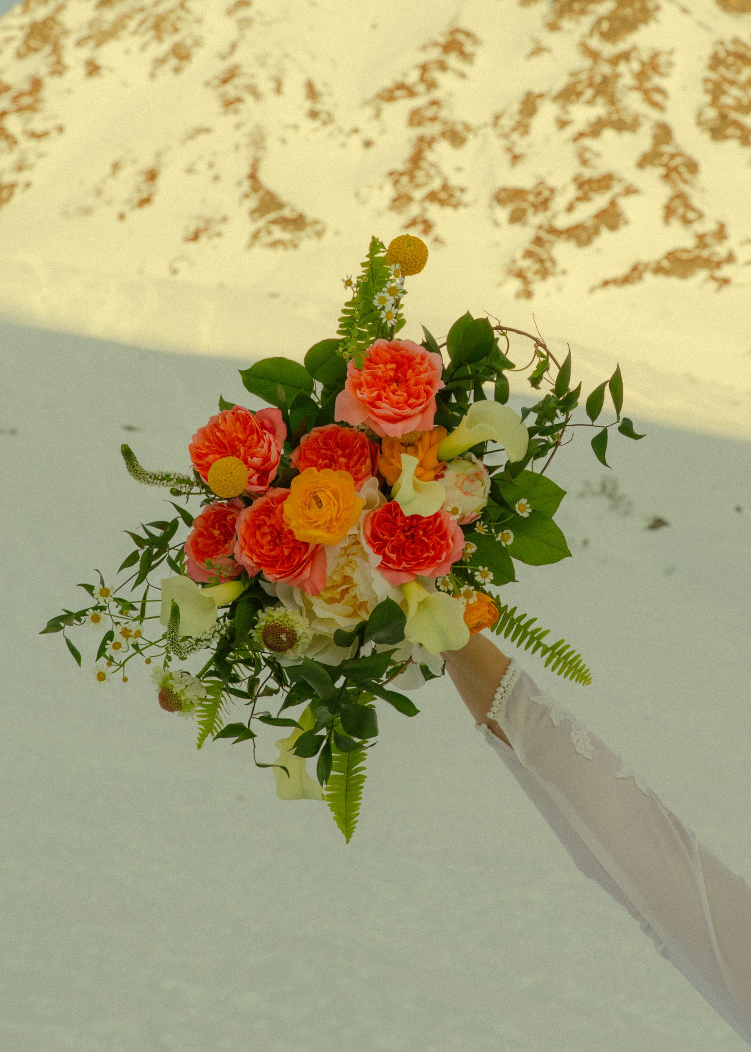 Elegant bridal bouquet with vibrant orange roses, green ferns, and yellow accents held against a snowy winter backdrop.