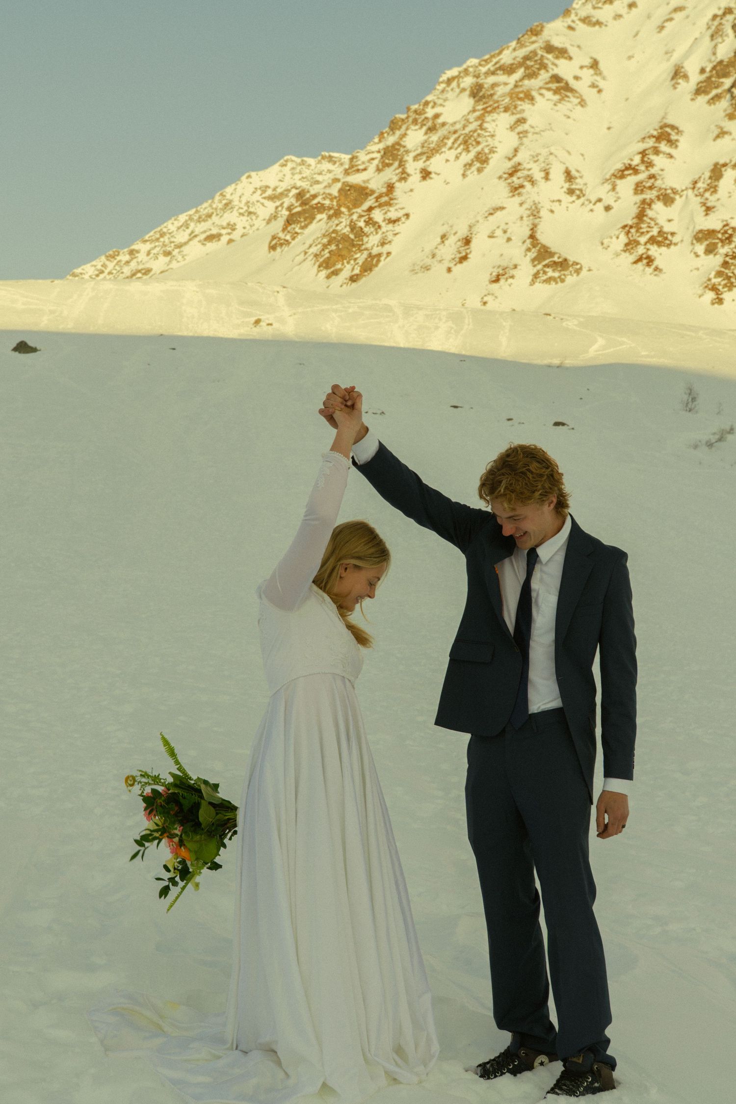 Groom spins bride in white dress on snowy mountain landscape while she holds a colorful floral bouquet.
