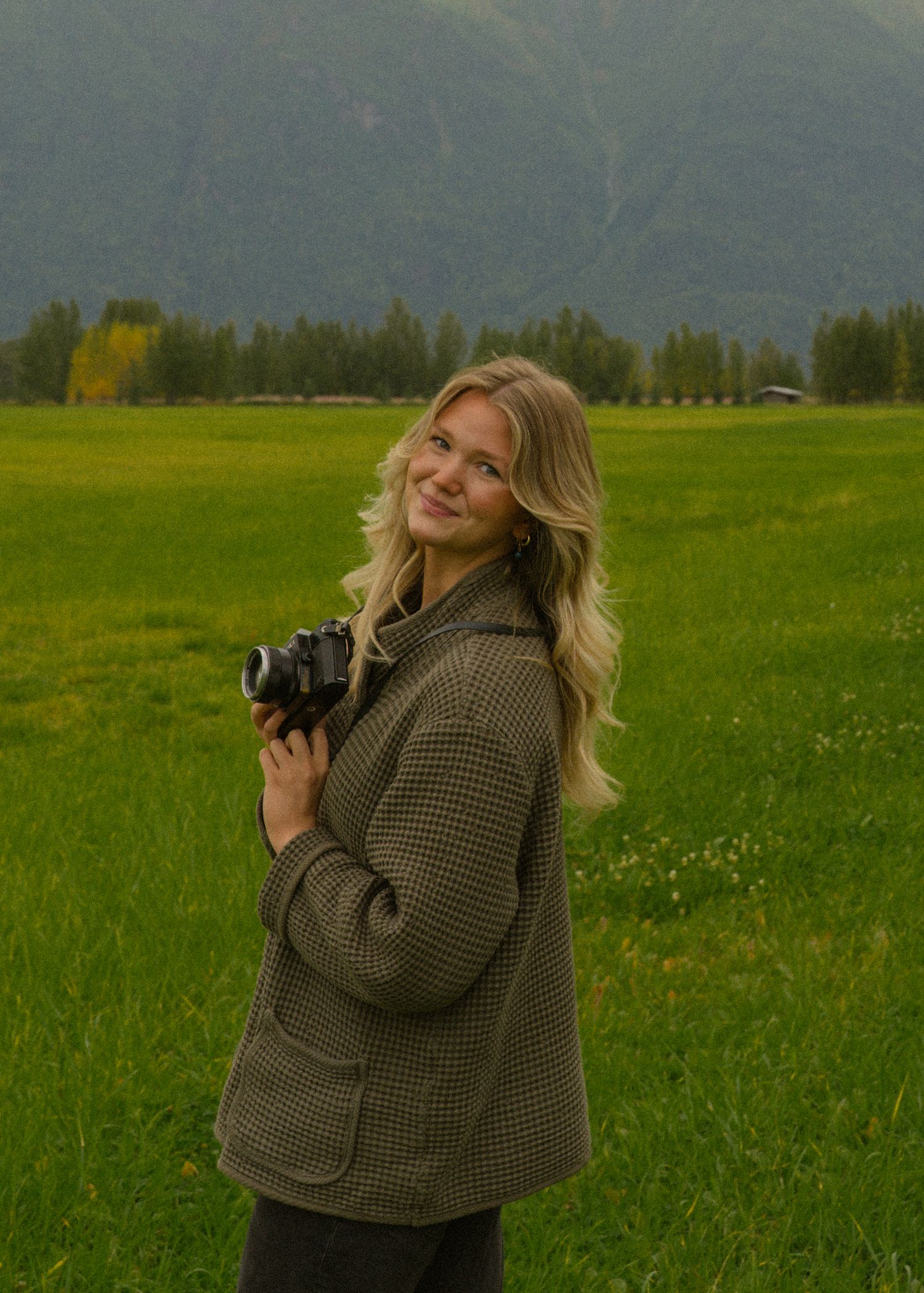 Smiling blonde woman holding a camera in a green field with mountains behind her.