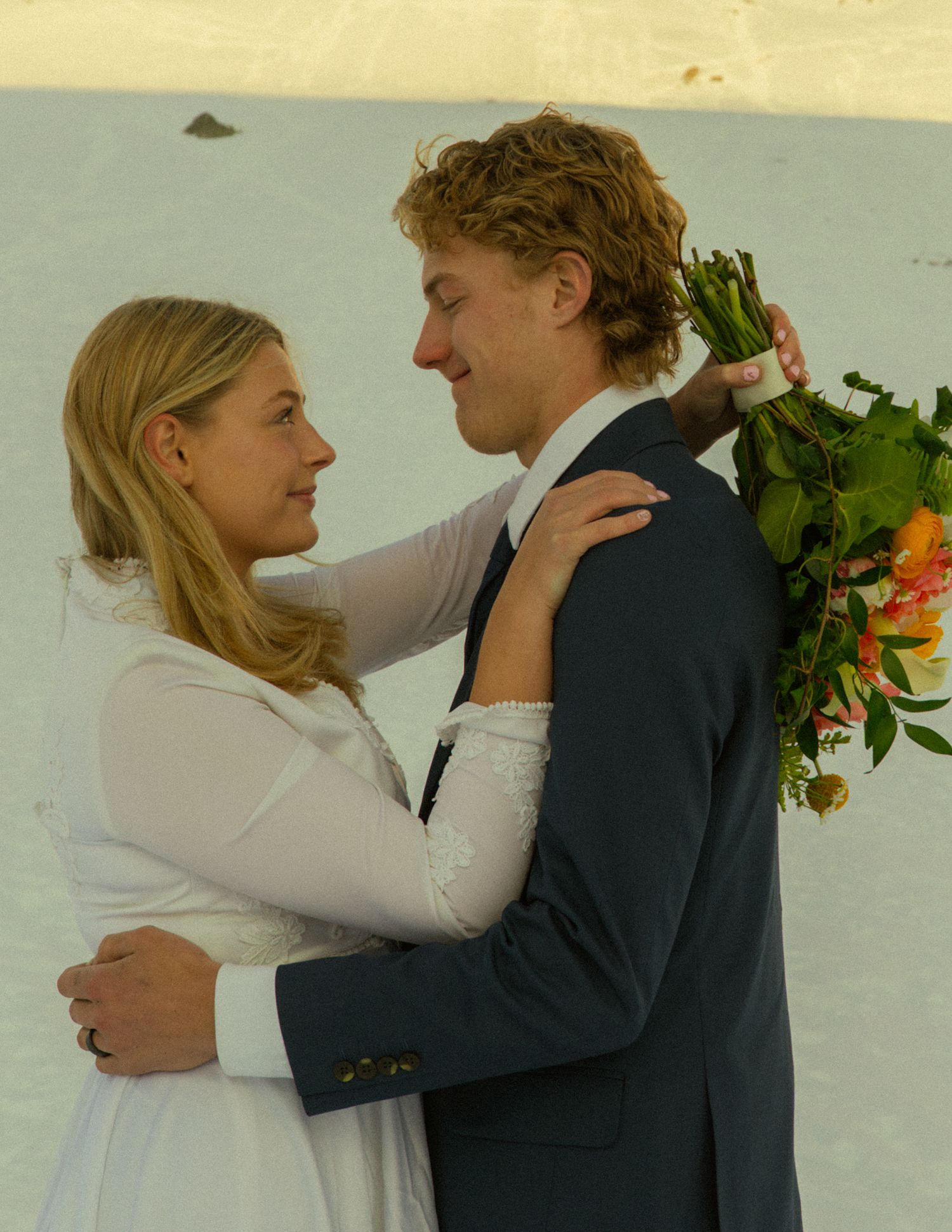 Smiling bride and groom in an intimate embrace in the snow, she in white dress, he in navy suit with floral bouquet.