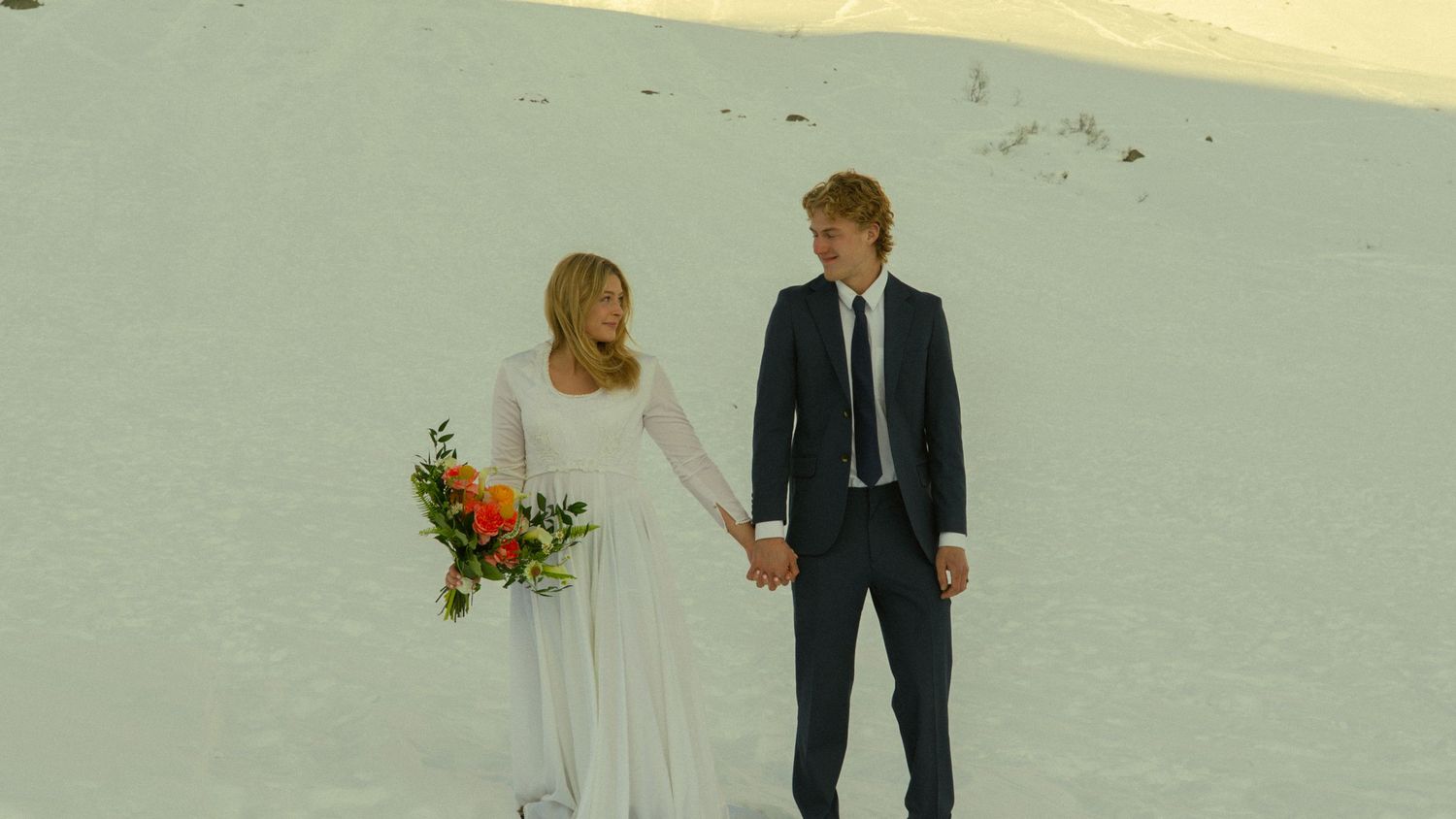 Newlyweds holding hands while walking across snowy landscape with mountain backdrop on wedding day.
