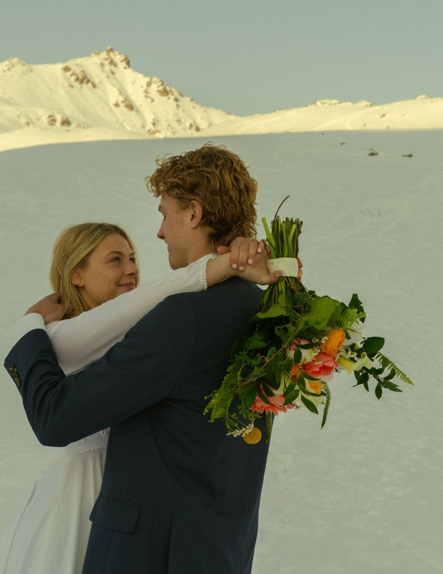 Couple embracing in snowy mountain landscape, bride in white dress holding colorful bouquet during winter wedding.