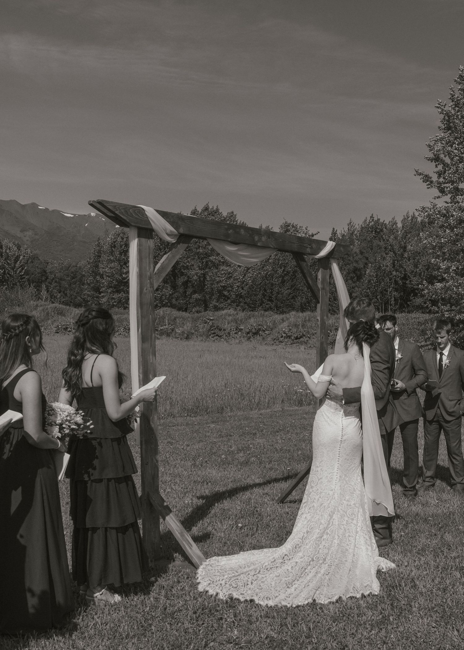 Black and white outdoor wedding ceremony with bride in lace gown at wooden arch, bridesmaids in dark dresses.