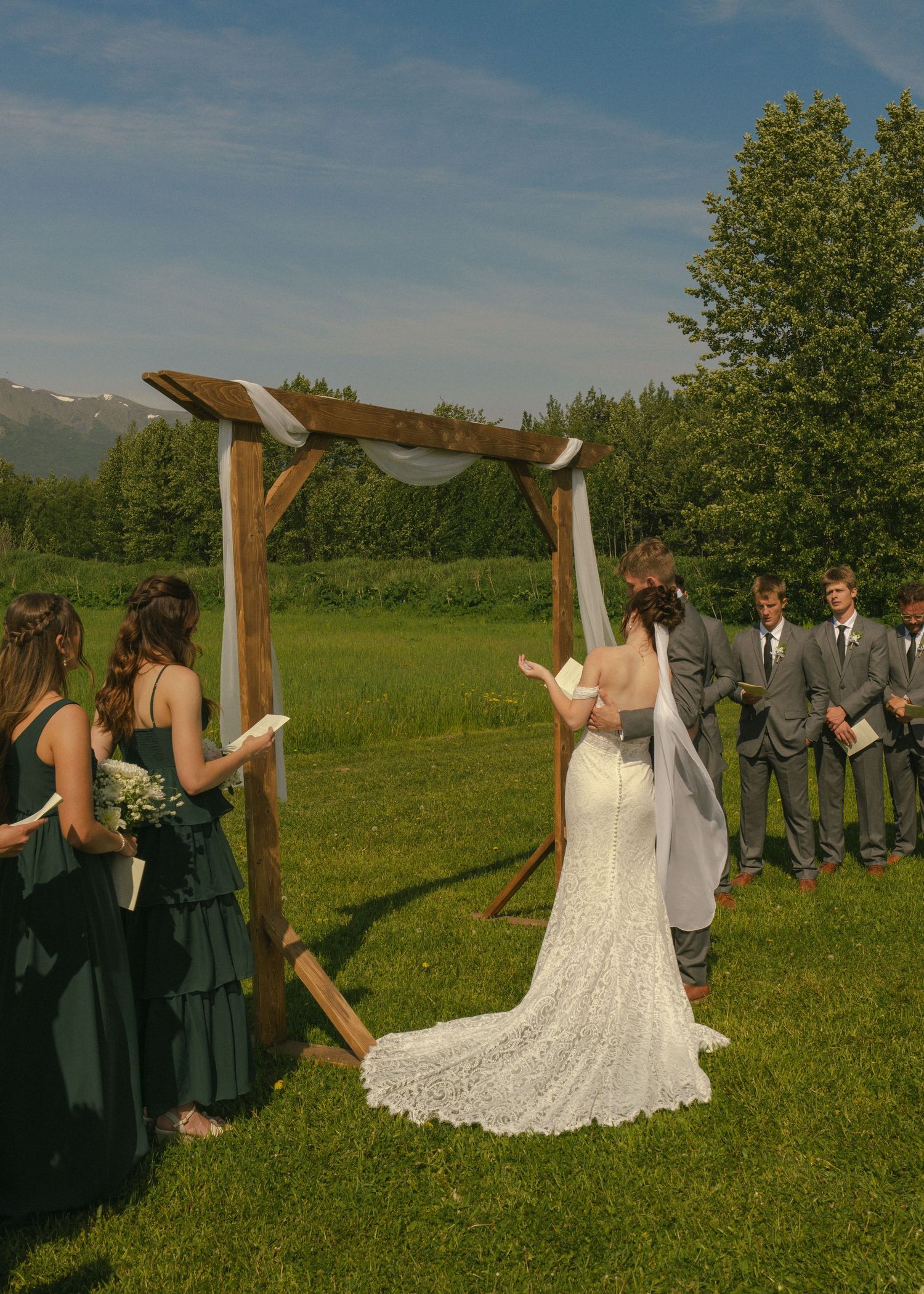 Outdoor wedding ceremony at rustic wooden arch with bride in lace gown, green-dressed bridesmaids, and groomsmen.