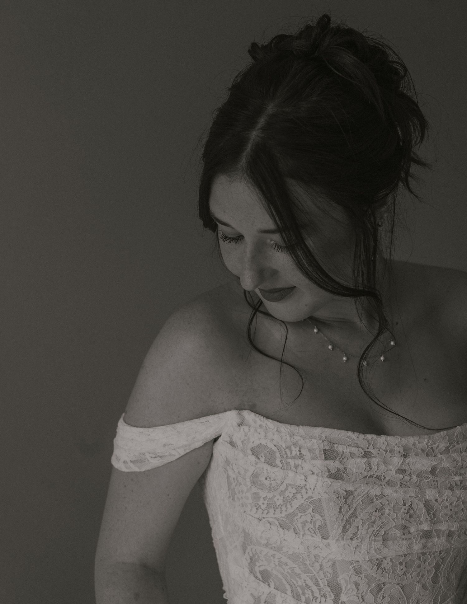 Black and white portrait of bride in off-shoulder lace gown with updo hairstyle and delicate necklace.