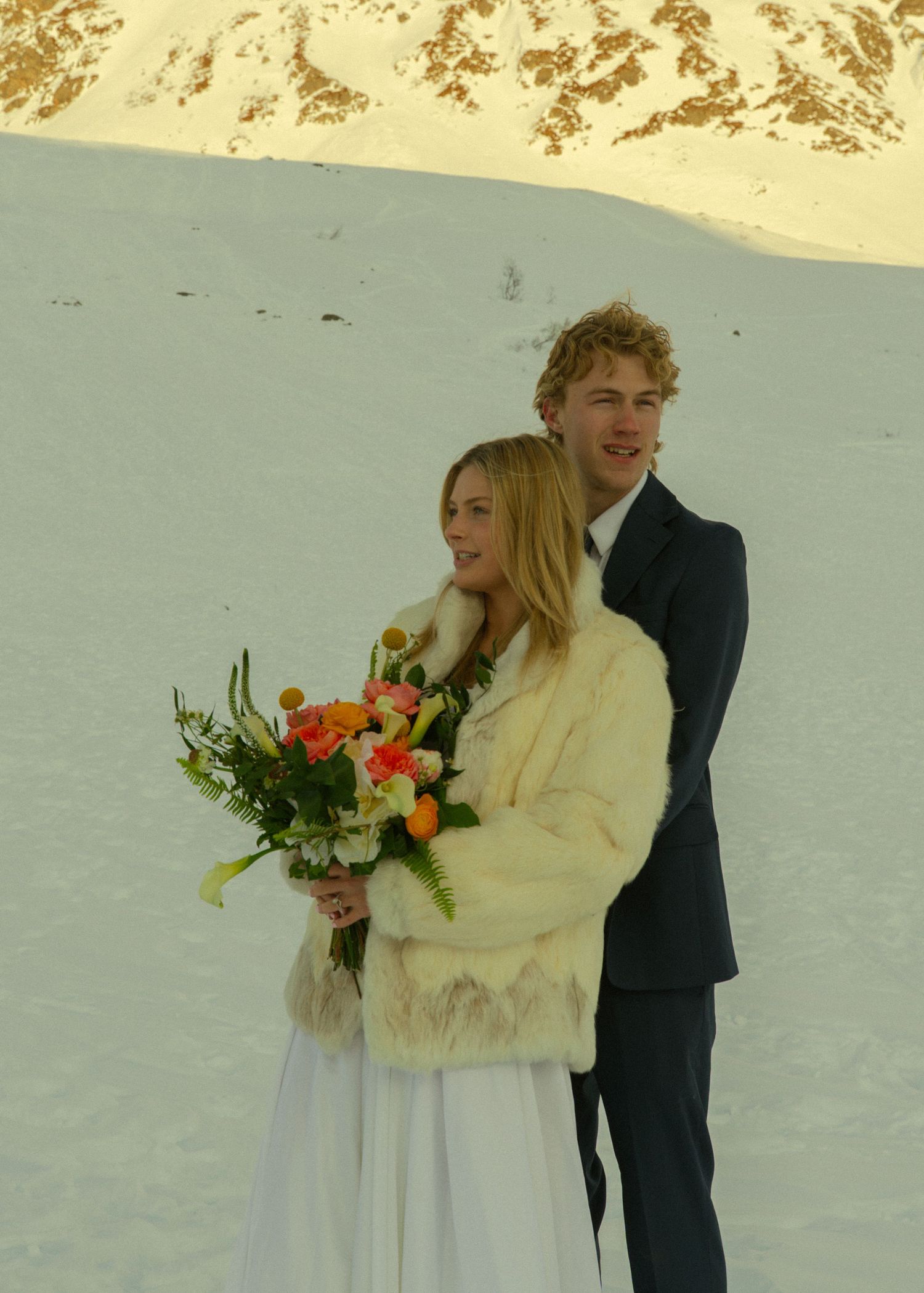 Bride in fur coat holding colorful bouquet with groom in dark suit posing on snowy mountain landscape.
