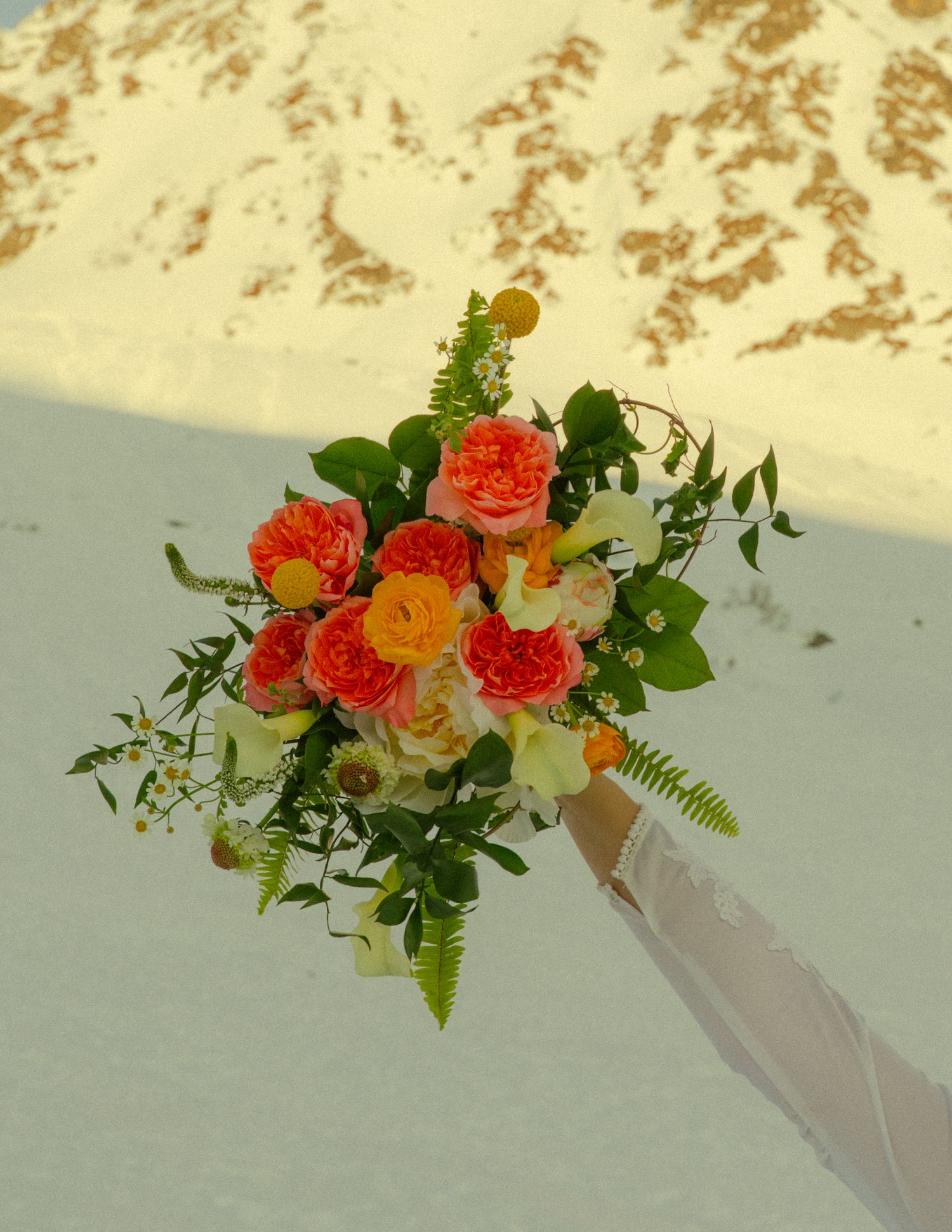 Close-up of a lush bridal bouquet featuring orange garden roses, ferns, and yellow blooms against a snowy mountain.