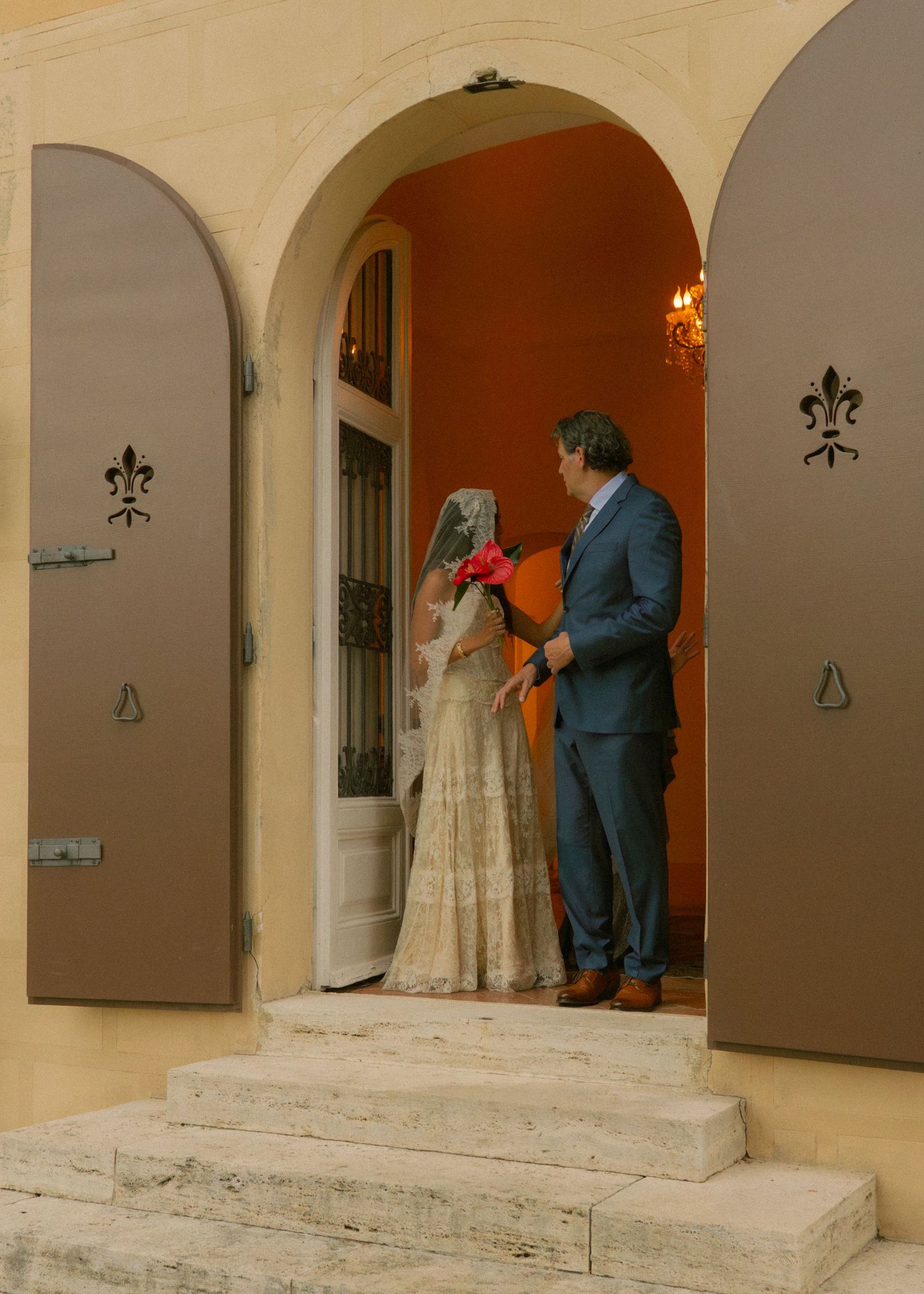 Bride in traditional attire with veil and groom in blue suit standing in ornate arched doorway with fleur-de-lis decor.