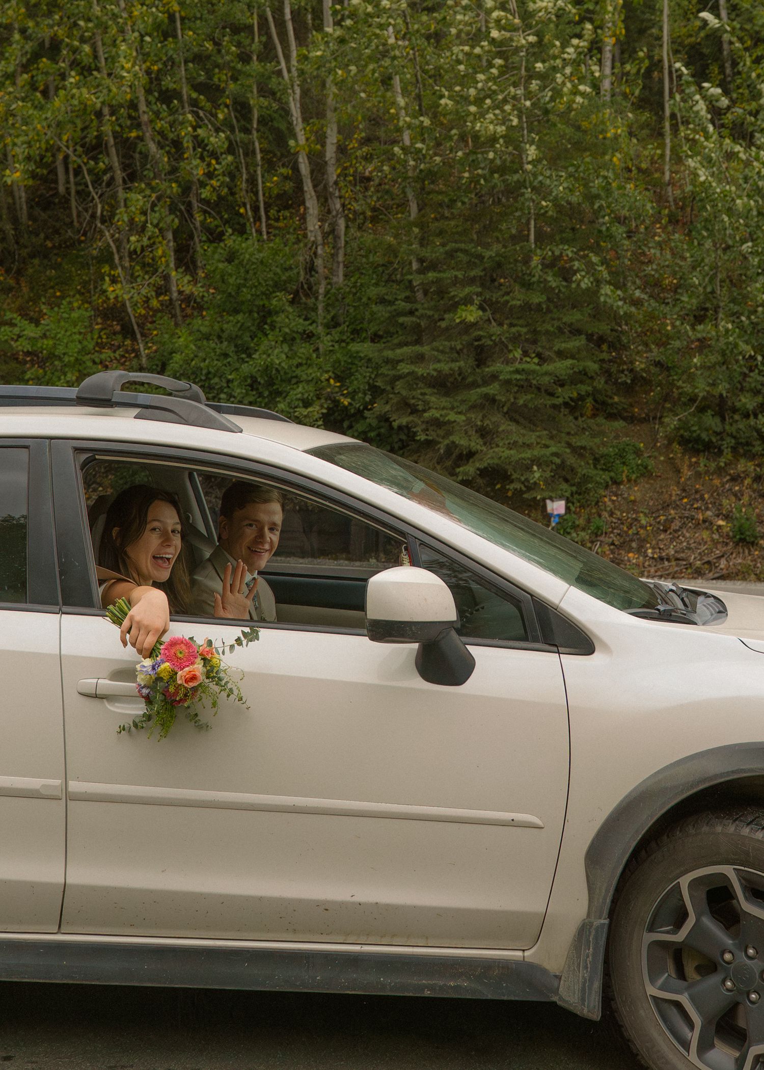 A newlywed couple smiles and waves from a white SUV, bride holding a colorful bouquet out the window.
