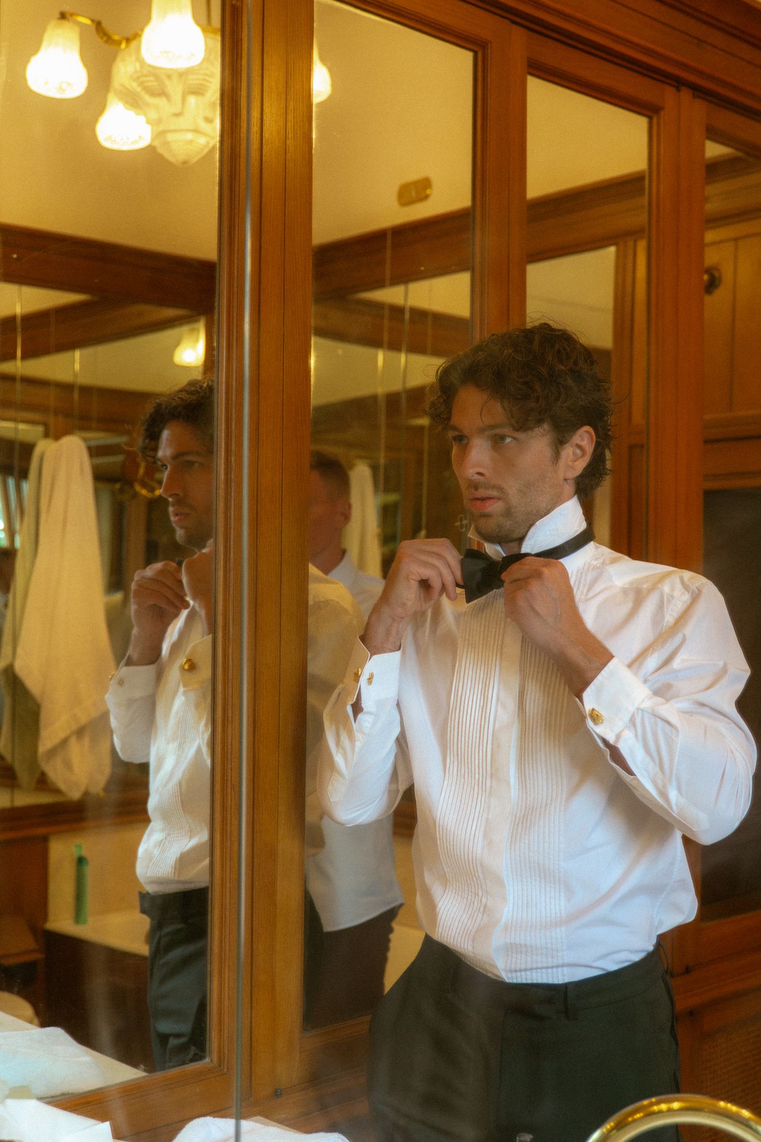 Groom in tuxedo shirt adjusting bow tie while looking in ornate wooden framed mirror before wedding.