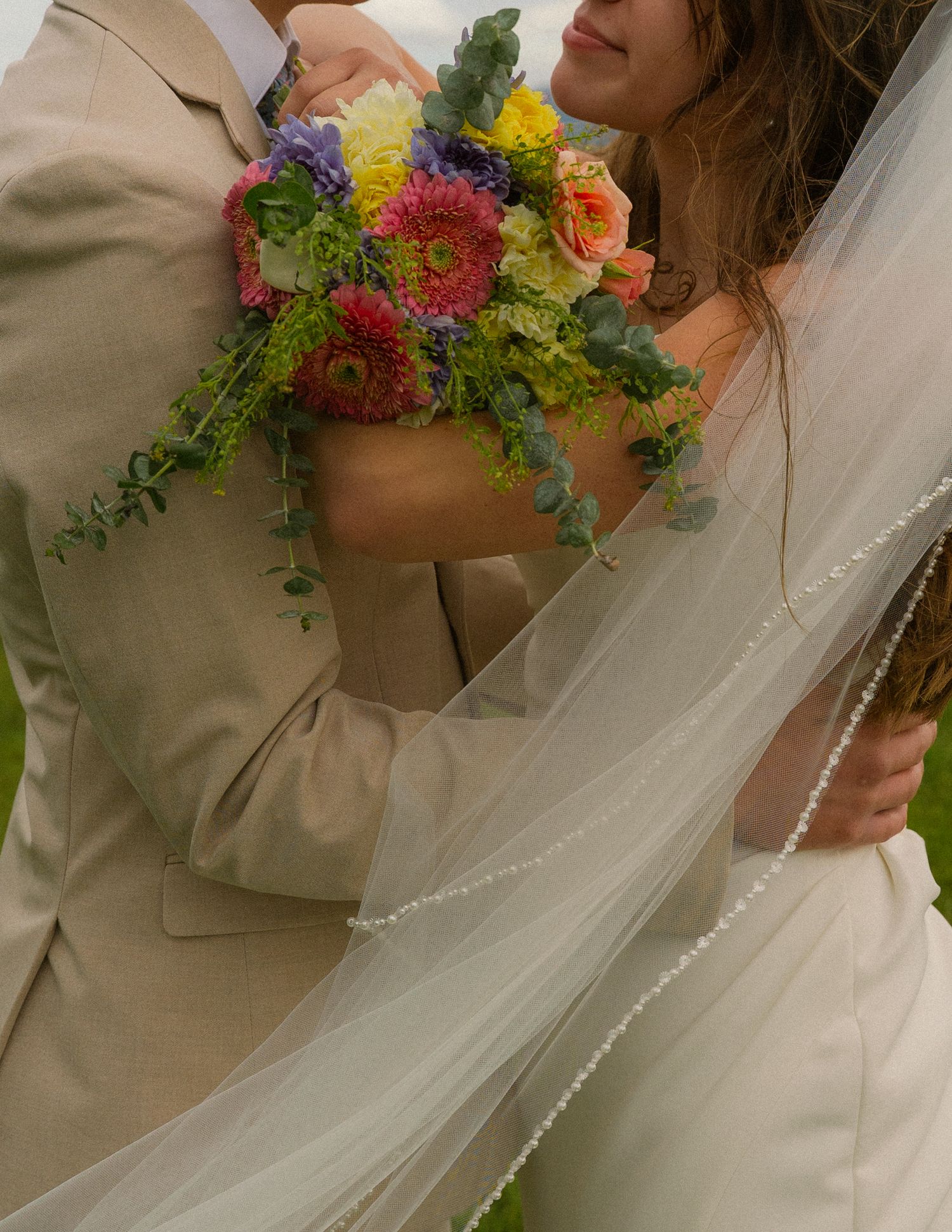 Bride and groom embracing, holding a colorful wedding bouquet with pink, yellow, and purple flowers, bride wearing a veil.