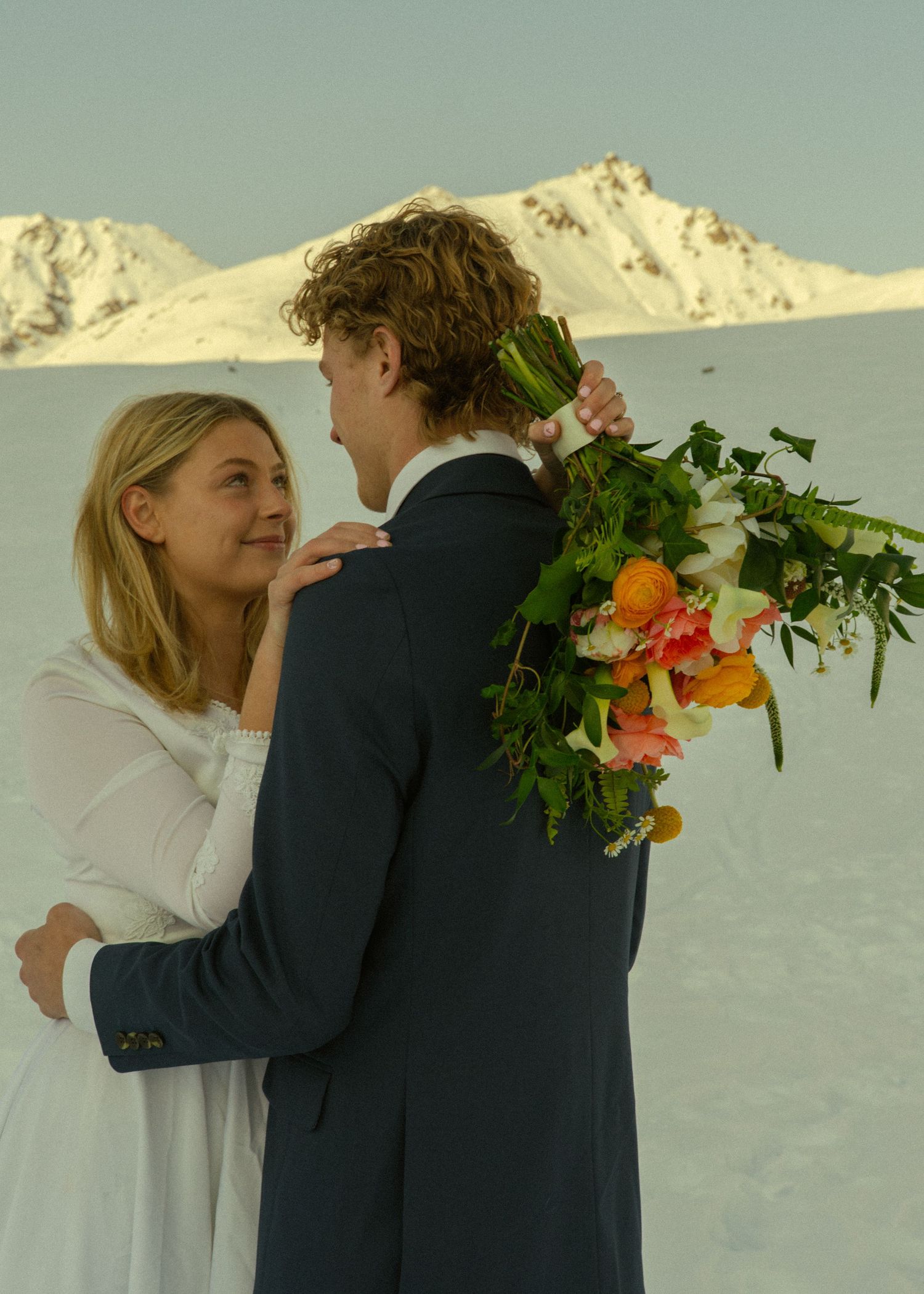 Bride in white dress embraces groom in navy suit as he holds a colorful bouquet against a snowy mountain backdrop.