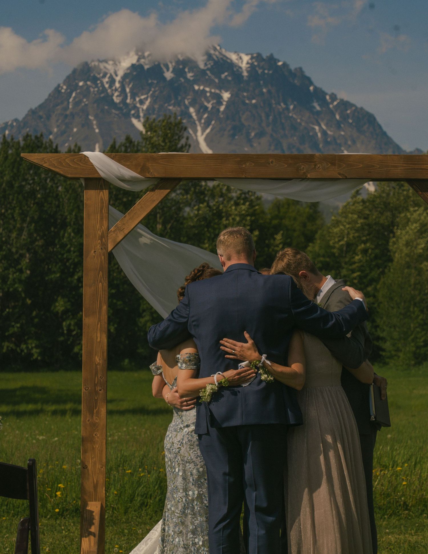Wedding ceremony under rustic wooden arch with snow-capped mountain backdrop as couple and guests share an embrace.