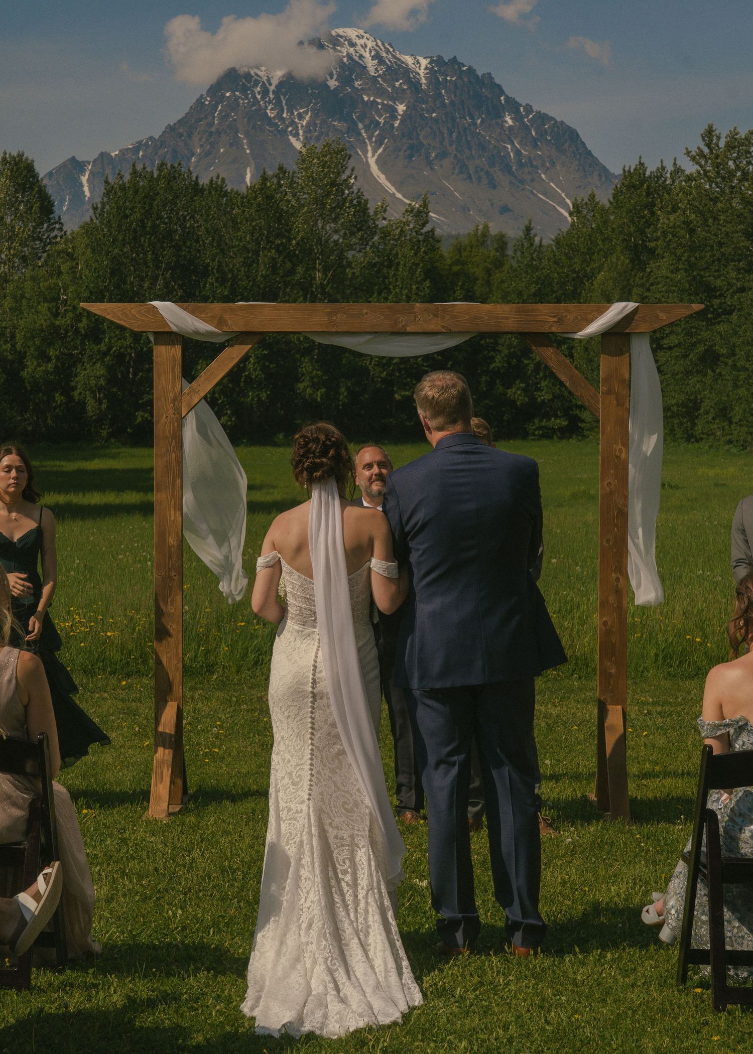 Bride and groom stand at rustic wooden arch during outdoor ceremony with snow-capped mountain backdrop.
