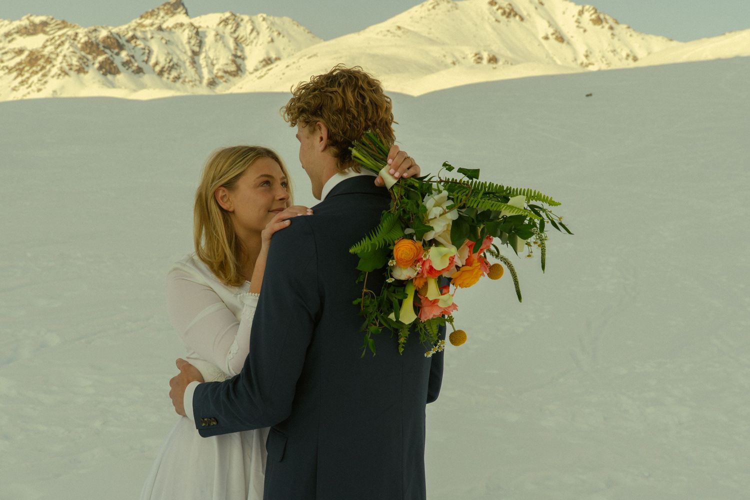 Bride and groom sharing intimate moment on snow-covered glacier with groom holding lush colorful bouquet.