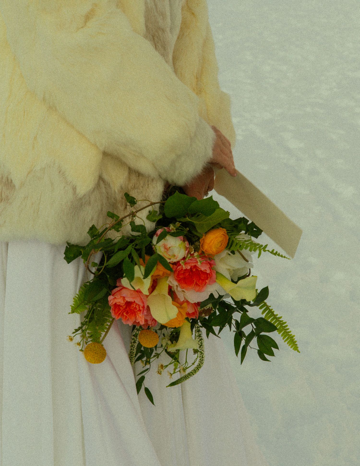 Bride in white gown and cream fur coat holding a colorful bouquet of orange roses and tropical flowers in the snow.