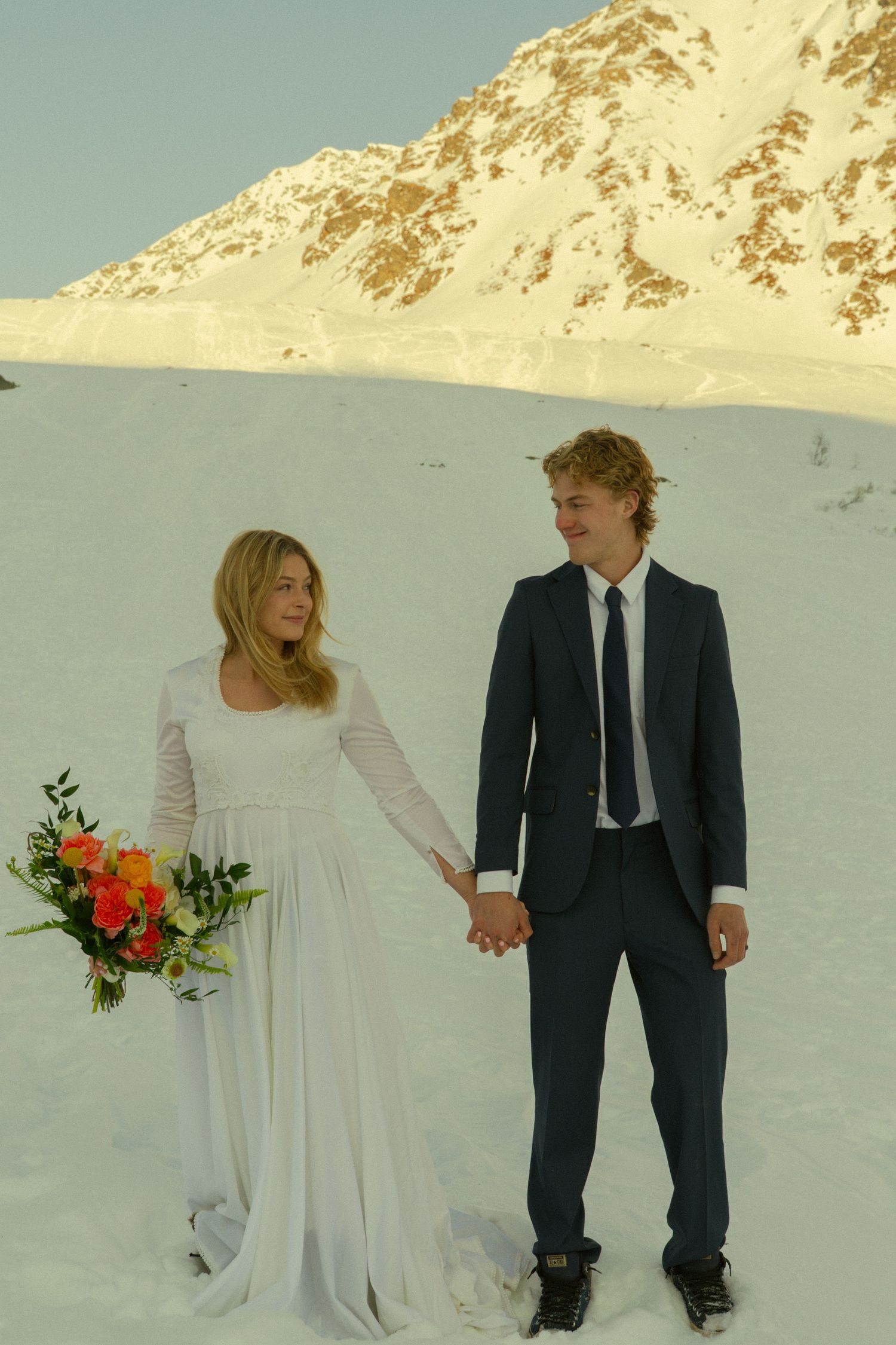 Bride and groom holding hands in white snow-covered mountain setting, bride carrying orange floral bouquet.