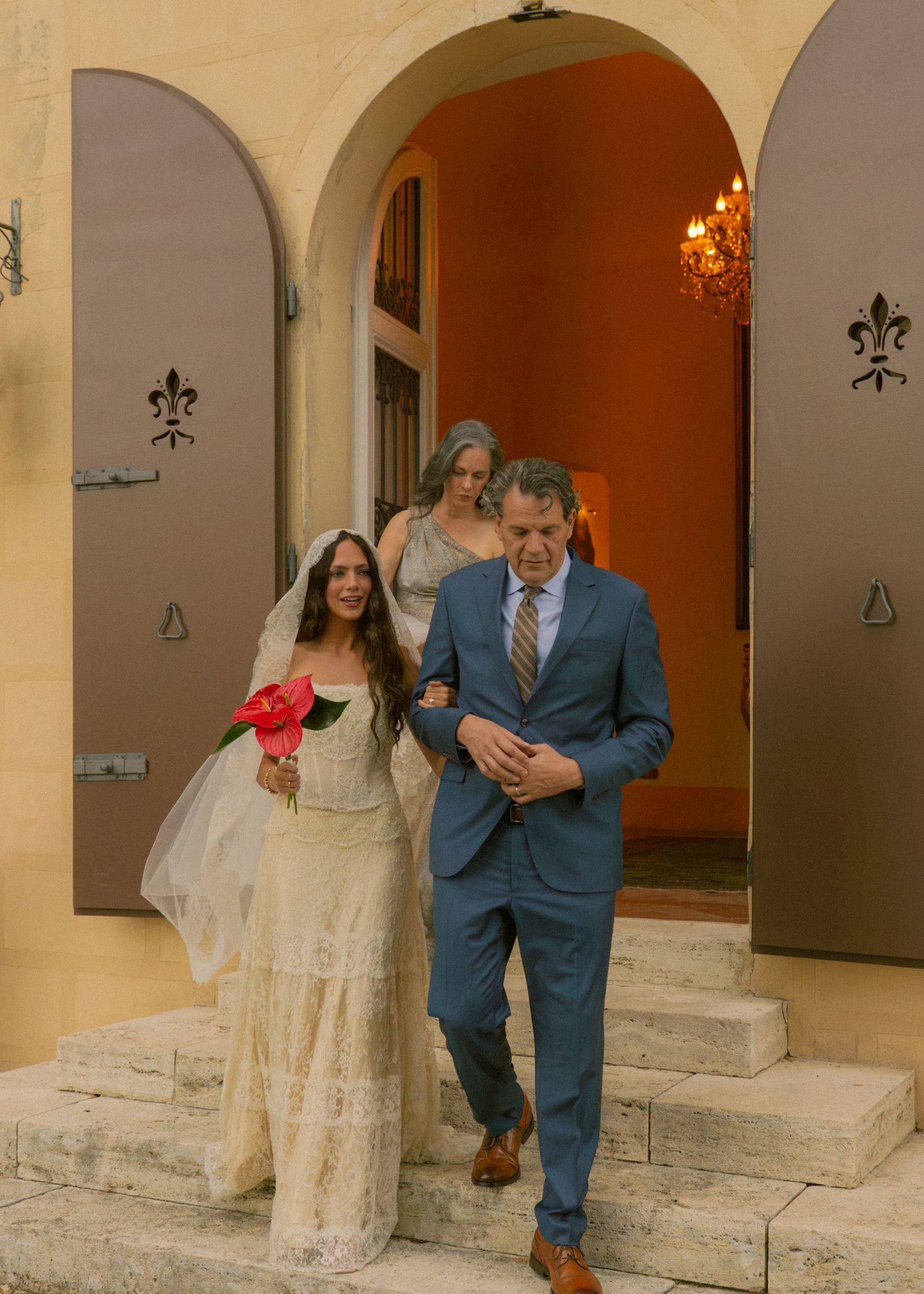 Bride in lace wedding gown holding red flower walks arm-in-arm with escort through ornate arched doorway.