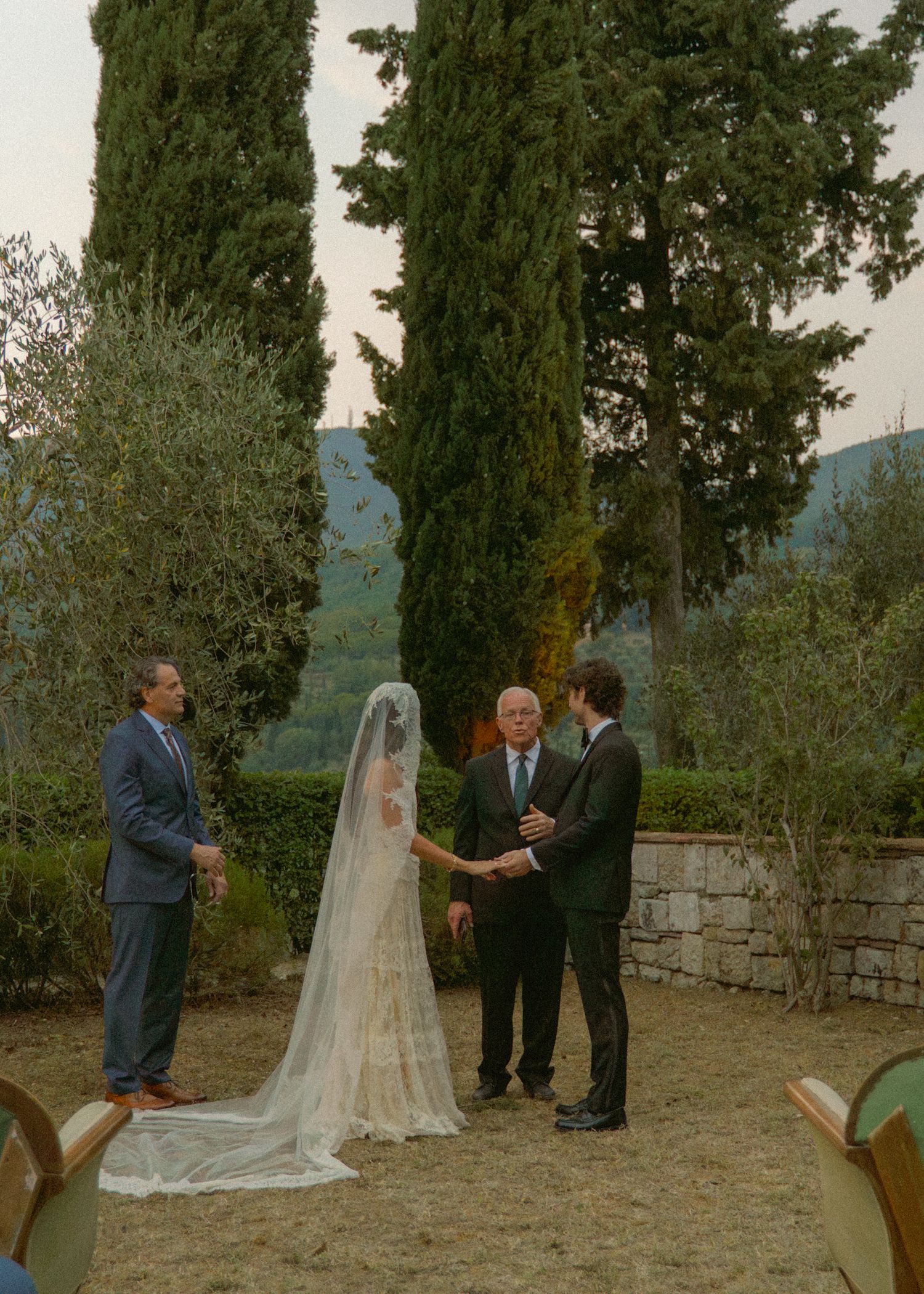 Wedding ceremony with bride in long veil and groom holding hands before an officiant outdoors.